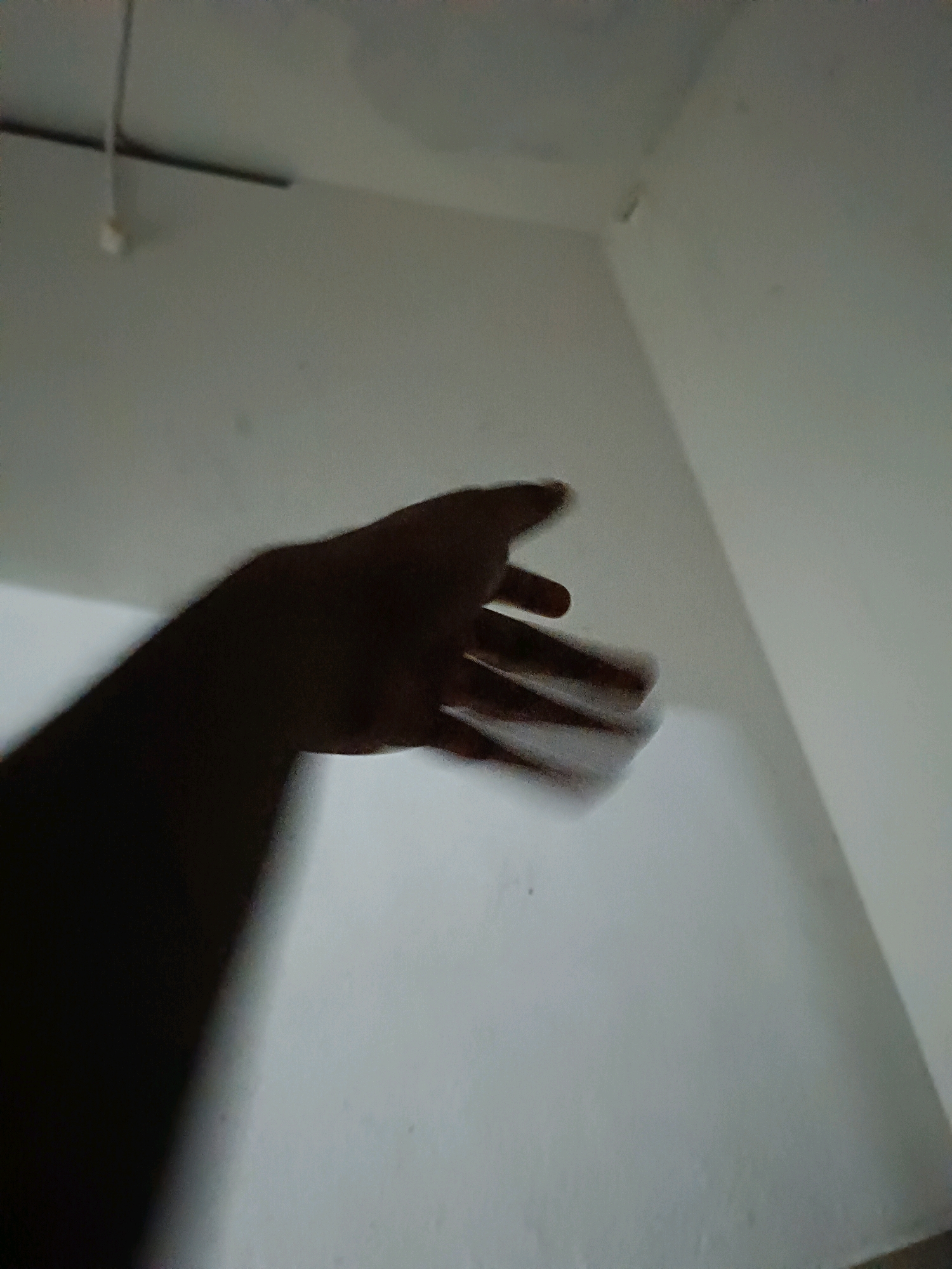 The image shows a dark shadow of a hand, with fingers extended and blurred due to motion, against a light-colored wall. The wall appears to be the interior of a room, possibly a corner where two walls meet. The lighting is dim, creating a strong contrast between the shadow and the wall. There is a thin, dark line extending horizontally across the upper left portion of the image, possibly a beam or shelf, with a small white object hanging from it by a thin string. The overall scene is abstract and lacks clear environmental context beyond the implied interior space. The location in Korani, India, is not discernible from the visual information presented in the image.