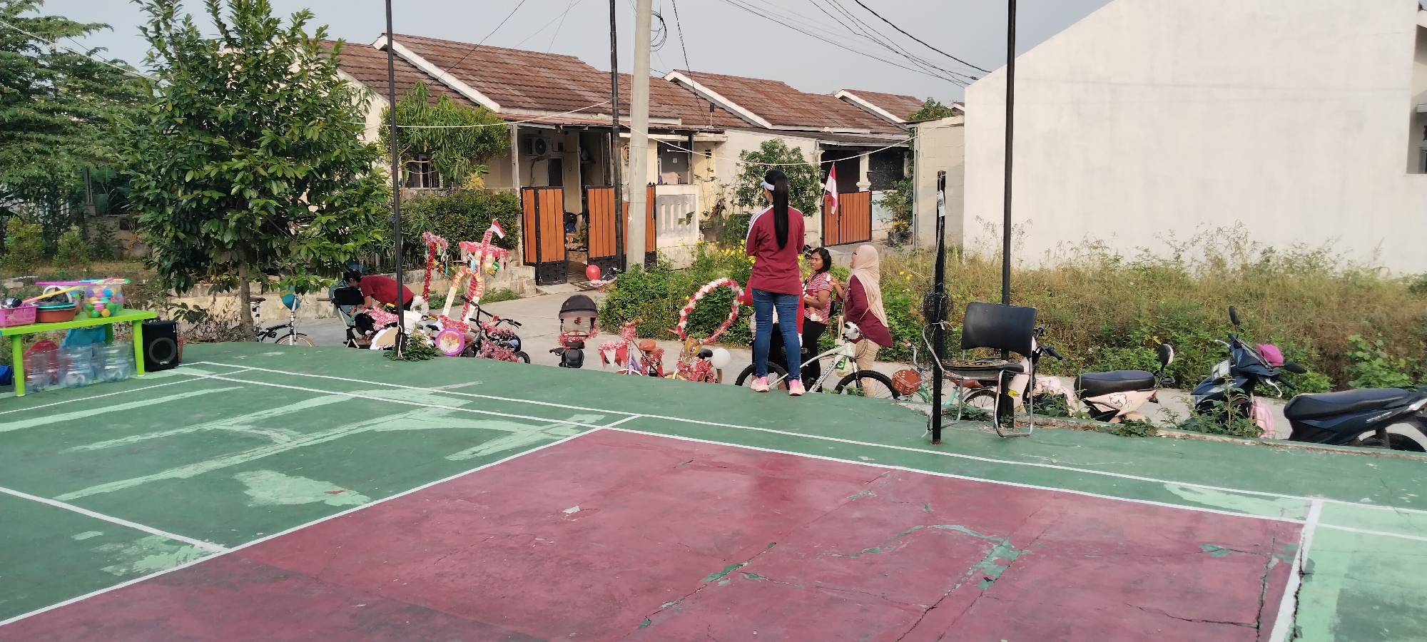 The image depicts a festive scene outdoors, likely in a residential area of Taban, Indonesia, possibly a neighborhood or housing complex. In the foreground, a partially visible badminton court with red and green surfaces is evident. To the left, a green table holds various items, including water bottles, colorful balls, and baskets. A black speaker is also placed on the ground near the table.
In the mid-ground, a line of decorated bicycles and what appear to be smaller celebratory floats are arranged. These decorations include red and white elements, ribbons, and possibly small flags, suggesting a national or local celebration. Several individuals are present, with three women in the center of the frame interacting. One woman in a red top and jeans stands facing away from the camera, while another woman in a hijab and a third woman in a striped shirt are beside her. Two individuals, possibly children, are bending over some of the decorated items. A bicycle and what looks like a stroller are also near this group. Further to the right, a black plastic chair sits near a pole, and several scooters are parked.
The background features a row of houses with brown tiled roofs and beige walls, typical of Indonesian residential architecture. Lush green trees and overgrown vegetation surround the houses and extend along the right side of the image. The sky is clear and bright, indicating daytime. The overall atmosphere suggests a community gathering or event.
