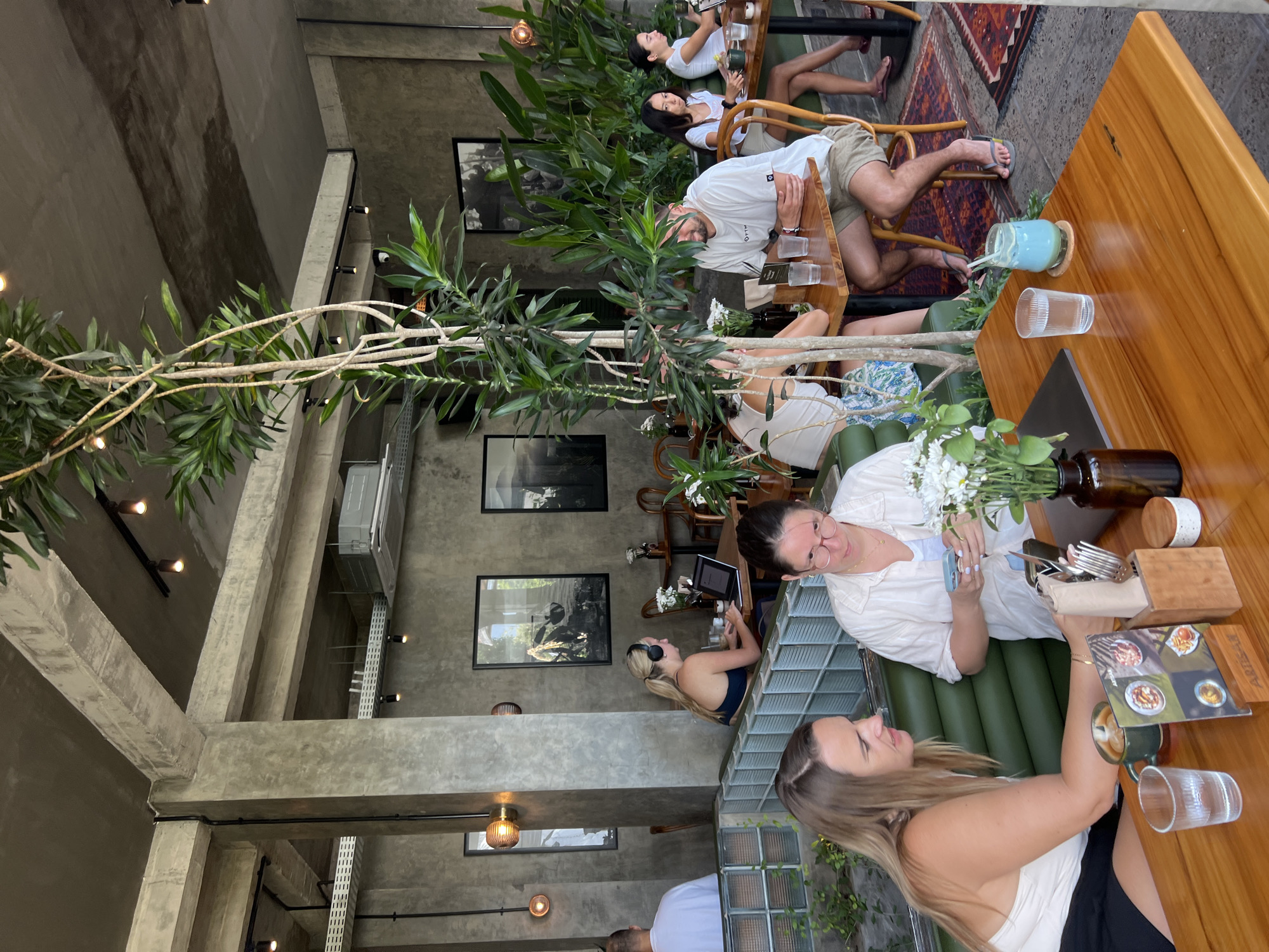 The image depicts the interior of a modern cafe in Mengwi, Indonesia, characterized by a raw concrete aesthetic, exposed beams, and abundant greenery. In the foreground, two women are seated at a wooden table. The woman on the left, wearing a white top and black shorts, leans forward, engaged with her phone. The woman on the right, with glasses and a light-colored shirt, is also looking at her phone. A menu, a cup of coffee, and a glass of water are visible on their table, along with a vase of white flowers.
Further back, a woman with blonde hair and headphones is working on a laptop at another table. Several other patrons are scattered throughout the cafe, some engaged with devices, others in conversation. The cafe's decor includes framed black and white photographs on the concrete walls, hanging pendant lights, and an industrial-style air conditioning unit. Lush potted plants and a trellis of vines add a natural element to the urban setting. The flooring transitions from polished concrete to a patterned rug near some of the tables. The overall atmosphere suggests a relaxed and contemporary dining or working environment.
