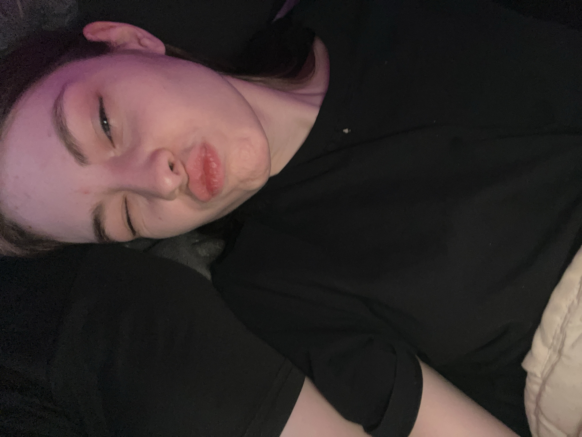 A pale-skinned person is lying down, with their head resting on a black cushion or surface. The person's face is turned towards the camera with closed lips, a slight pout, and their eyes partially closed. The individual is wearing a black t-shirt. The background is dark and indistinct. No indication of the location of Saint Petersburg is included in the image.