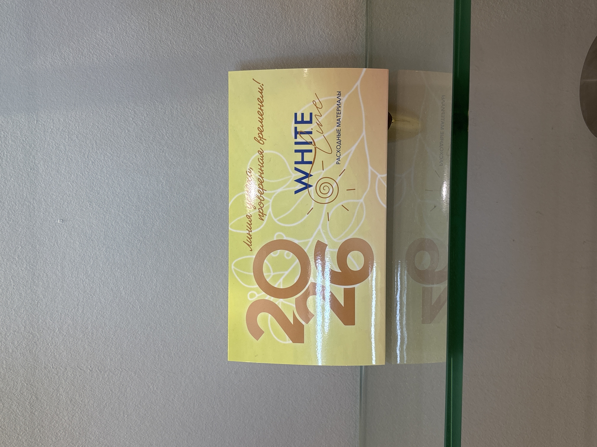 The image shows a close-up of a yellow and white label with Russian text and a graphic. The label is attached to a surface that appears to be a glass shelf or partition, indicated by a vertical green edge on the right side of the frame.
The subjects on the label include stylized text and graphic elements. There are large, orange numbers "20" and "26" prominently displayed, possibly indicating a year or a quantity. Above these numbers, in a script font, is the text "Лица, ухода, проверенная временем!" which translates to "Faces, care, time-tested!". Centered on the label is the brand name "WHITE line" written in a stylized font with the word "line" in a script. To the right of the brand name, in smaller capitalized letters, is "РАСХОДНЫЕ МАТЕРИАЛЫ", meaning "CONSUMABLES". The graphic elements consist of a circular design resembling a sun or a swirl, with radiating lines, and abstract leaf-like patterns.
The scene appears to be an indoor setting, possibly a retail environment or a display area, given the presence of the glass and the nature of the label which suggests a product or promotional item. The lighting is even, and there are no obvious indicators of the time of day or weather. The overall presentation is clean and visually organized, with a focus on the product information and branding. There are no people visible in the image.