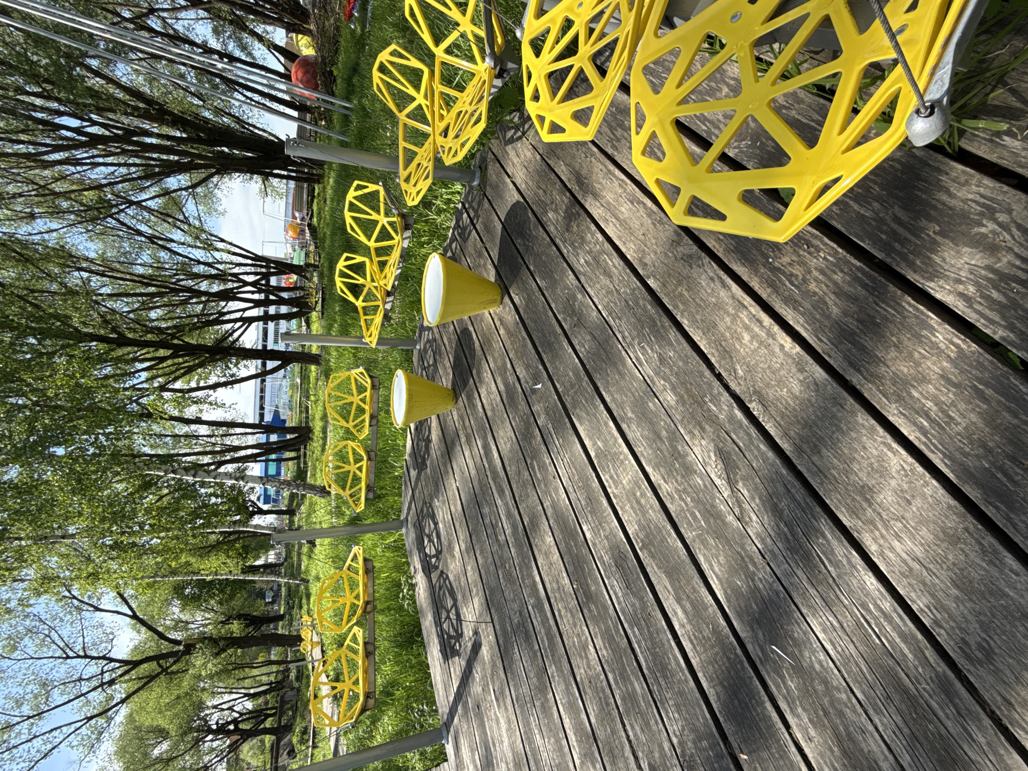 On a sunny day in Синьково, Russia, the scene is dominated by a wooden boardwalk extending diagonally across the frame from the lower right to the upper left. Along the edge of the boardwalk, near the upper left corner, there is a row of yellow geometric sculptures mounted on metal poles, creating an interesting linear pattern against the backdrop of trees and a distant body of water.
The wooden planks of the boardwalk are weathered and gray, showing the grain and texture of the wood, with varying shades of light and shadow. The yellow sculptures have a complex, angular design, resembling cages or abstract forms. Some of the sculptures are cone-shaped at the bottom with a white interior.
Behind the sculptures, trees with green foliage can be seen, and in the distance, there is a body of water, suggesting a lake or river. The sky is visible through the branches of the trees, hinting at a bright day. The photograph appears to be taken from a low angle, emphasizing the perspective of the boardwalk and the sculptures.
