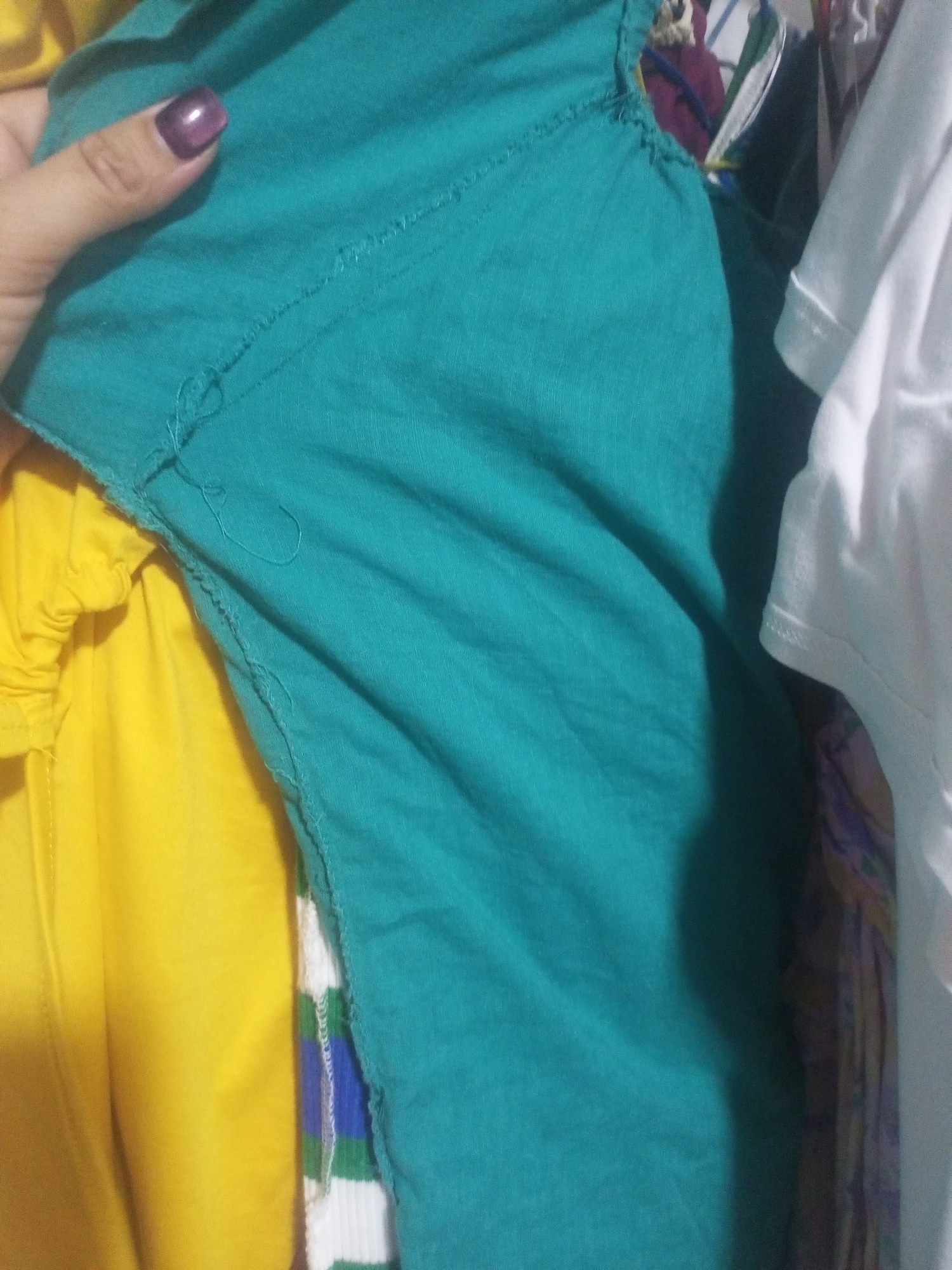 This is a close-up, waist-up photograph of a person's hand holding a teal-colored garment. The hand is positioned in the upper left quadrant of the image, with a dark purple nail polish visible on the fingernail. The garment appears to be made of a lightweight fabric and has a slightly frayed edge along the bottom. To the left of the teal garment, a bright yellow fabric is visible, possibly another piece of clothing. To the right of the teal garment, a white shirt sleeve with a ribbed cuff is visible, and behind that, a patterned fabric can be seen. The overall lighting suggests an indoor setting, with shadows cast on the teal fabric. There are no discernible background elements that would indicate a specific location in La Tagua, Venezuela, or any other city.