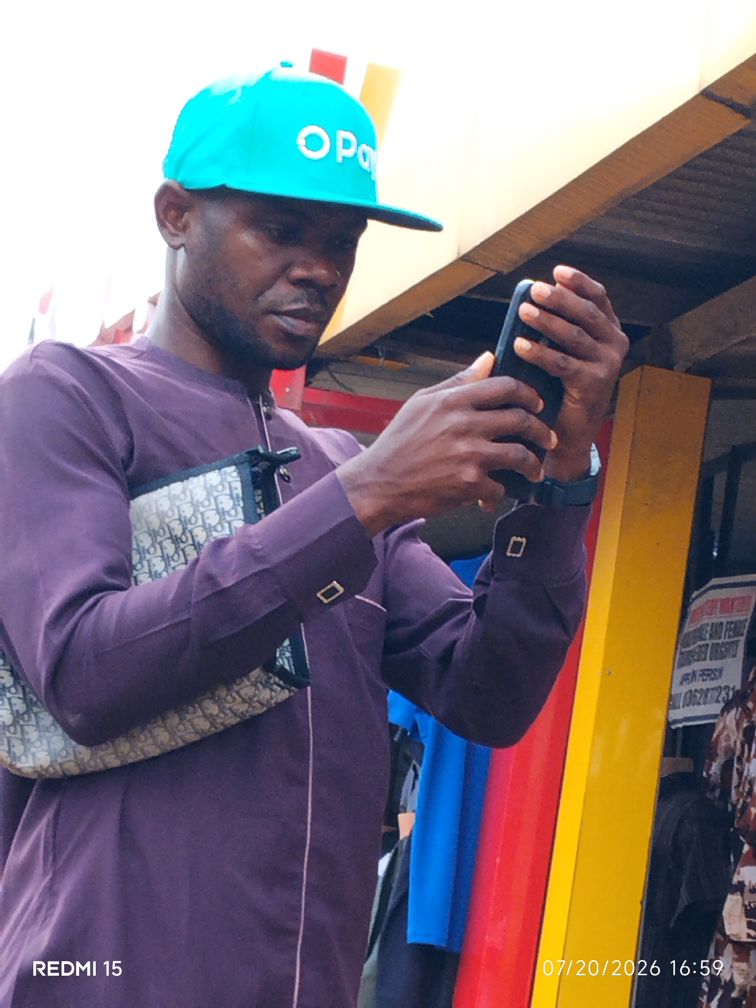The image captures a medium shot of a Black man in Umuode, Nigeria, wearing a purple traditional outfit and a bright turquoise cap with the "Opa" logo. He is looking down at his black smartphone, which he holds in his hands. The man is also carrying a patterned handbag over his left arm. In the background, vertical elements of red, blue, and yellow are visible, possibly part of a market stall or structure. A sign with text and a phone number is partially visible on the right. The timestamp "07/20/2026 16:59" and "REDMI 15" are overlaid on the image.