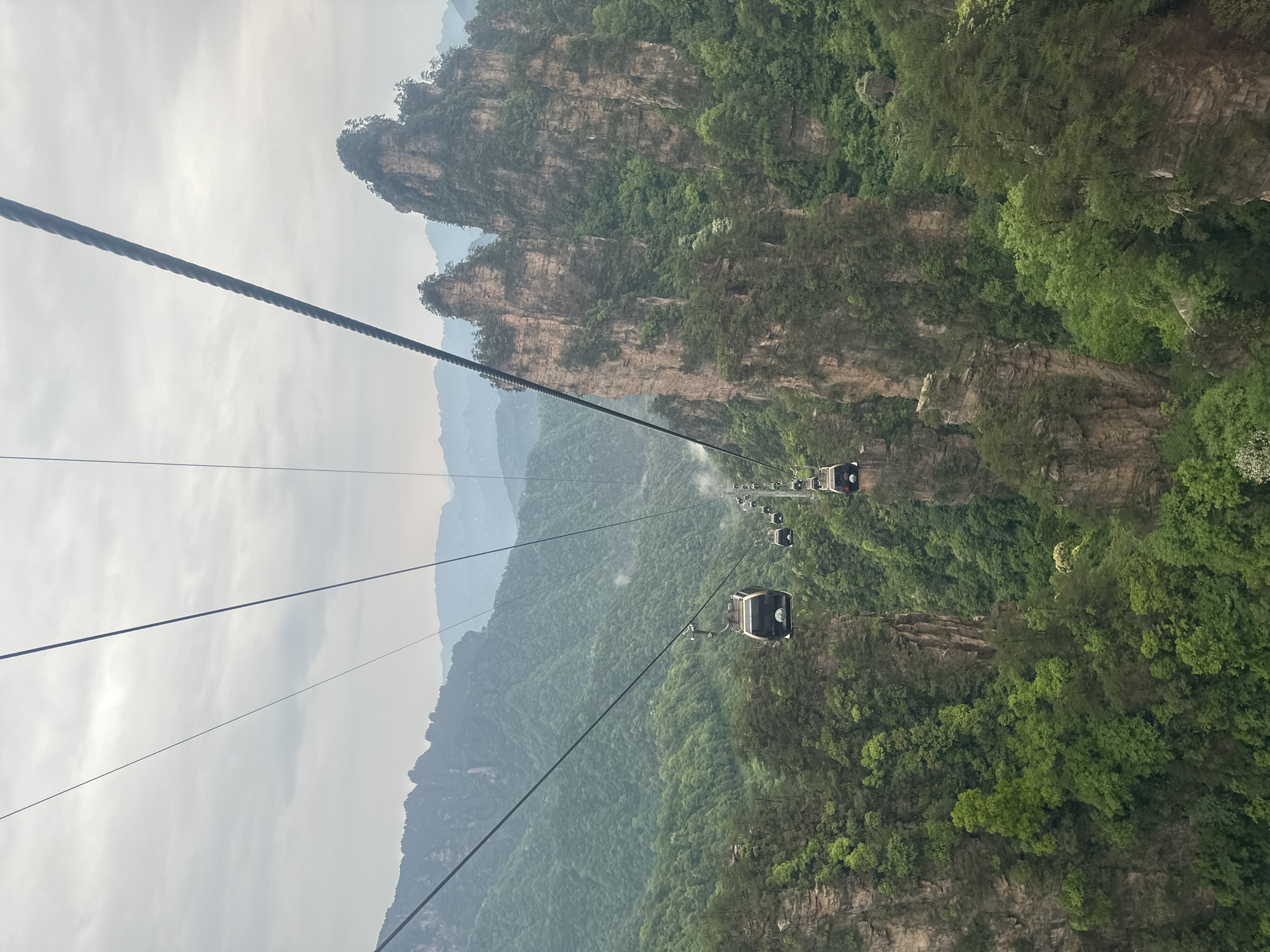 The image depicts a cable car system traversing the Tianzi Mountain in Wulingyuan, China. The cable cars are suspended over a landscape of densely forested mountains and towering rock formations. The sky is overcast and a light haze hangs in the air. The photograph is taken from within a cable car looking out at the descending cable cars.