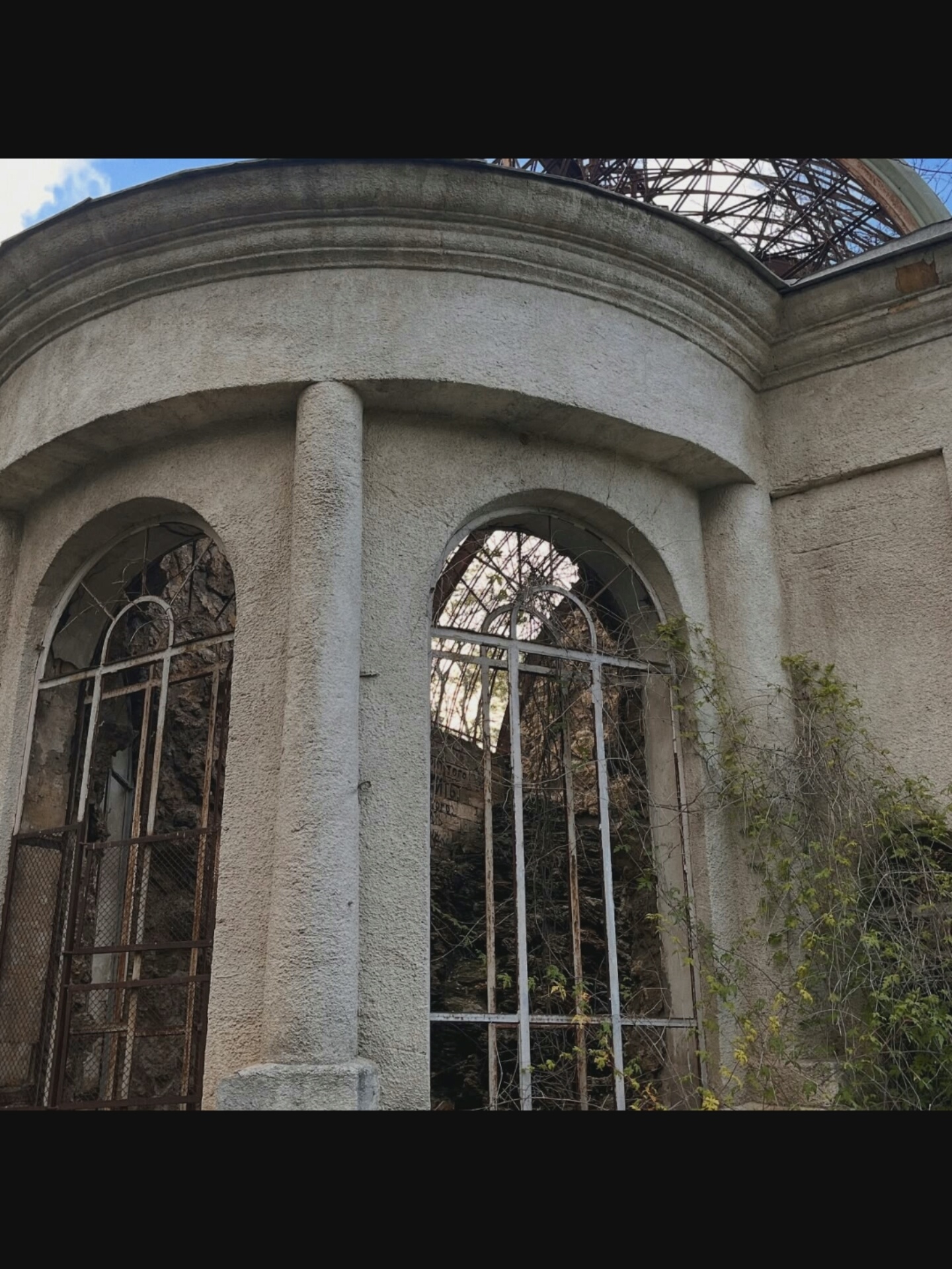 The image shows the exterior of a derelict building with arched windows. The building's facade is textured and light-colored, with a decorative cornice at the top. Two large arched windows are visible, each covered by metal grating. The windows are partially obscured by vines and overgrowth. Through the grating, it is possible to see the rough stone interior of the building, suggesting it is in a state of disrepair. Above the facade, the skeletal remains of a dome structure are visible against a cloudy sky. The overall impression is one of abandonment and decay. The location is identified as Bielawska Wieś, Poland.