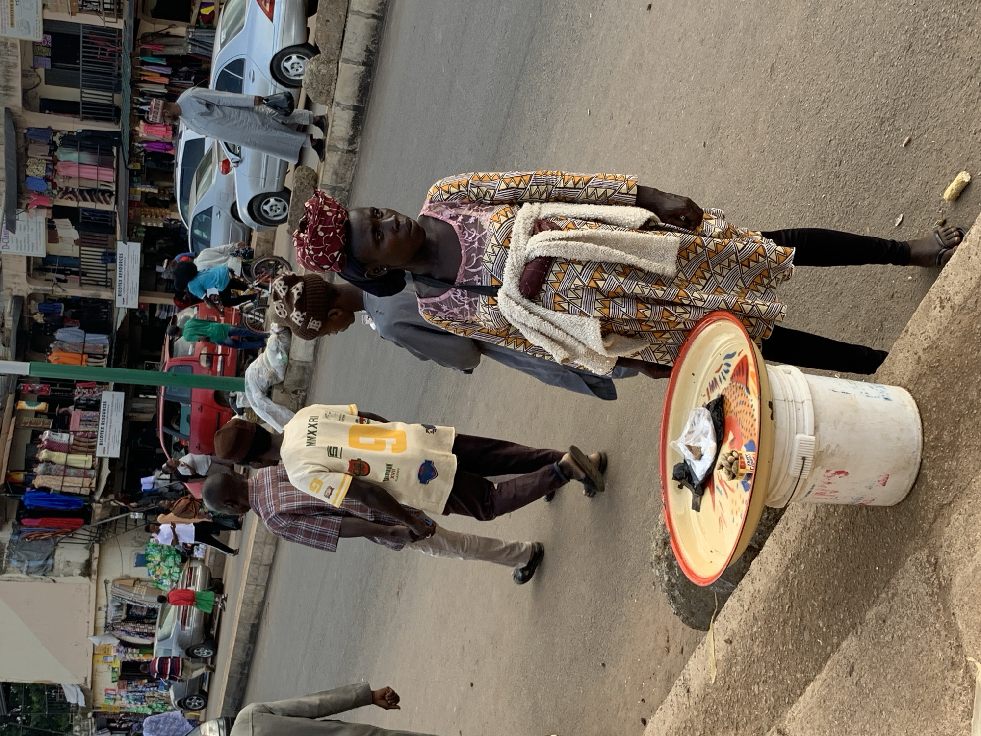 In Jos, Nigeria, a street scene unfolds with a woman prominently featured in the foreground, selling goods from a bucket topped with a decorative tray. The tray, with a red rim, is adorned with a bird motif and holds a plastic bag of items and a small container of nuts. The woman, wearing a vibrant, patterned dress and black leggings, stands by the roadside. Behind her, several other individuals are visible, including a man in a light-colored robe and a cap, and others walking along the street. The background reveals a bustling market environment with shops displaying clothing and various goods. Vehicles, including cars and a red car, are parked or moving along the street. The scene is set on a paved road with a sidewalk and curb.