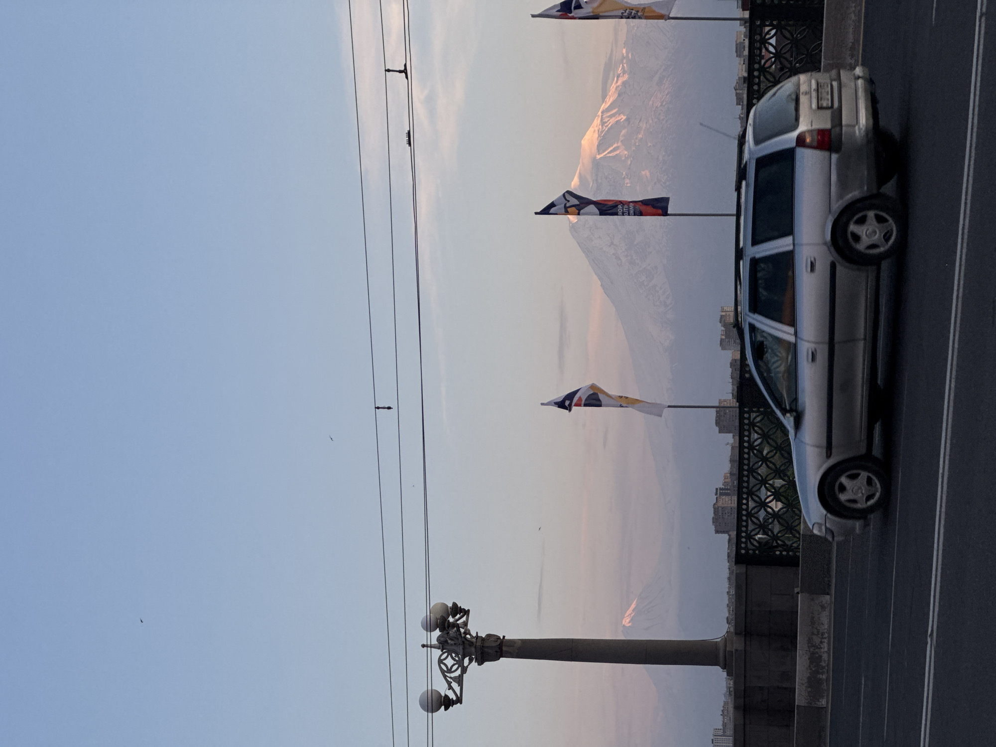 A serene urban landscape is captured during either sunrise or sunset, featuring a majestic, snow-capped Mount Ararat in the background, identifying the location as Yerevan, Armenia.
In the foreground on the right, a silver station wagon, possibly an Opel Zafira, is parked or moving slowly on a paved road, which appears to be part of a bridge due to a decorative metal railing. To the left, a tall, ornate lamppost with multiple lights stands prominently. Several power lines stretch across the upper left section of the pale blue sky.
Three vertical flags are unfurled by a gentle breeze. The middle flag clearly displays the word "EUROPEAN," possibly followed by "COMMUNITIES" or "CULTURE," and another flag features a distinct penguin-like character. Distant city buildings are visible between the road level and the base of the mountain.
The sky transitions from a light blue at the top to a soft, pale pink and orange gradient towards the horizon, casting a warm glow on the distant mountain peak. Small, dark specks indicating birds in flight can be seen against the sky. The overall atmosphere is peaceful, with clear weather and excellent visibility of the iconic mountain.