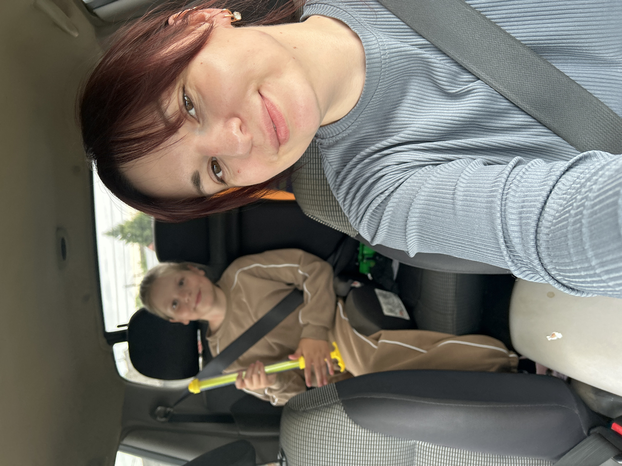 In the backseat of a vehicle, a woman with reddish-brown hair and bangs smiles at the camera, wearing a blue ribbed shirt and a gray seatbelt. To her left, a young girl with blonde hair, wearing a tan tracksuit, sits in a car seat and holds a yellow toy gun. The interior of the car is visible, with patterned seats and car seat components. Outside the car windows, trees and a cloudy sky can be seen, suggesting an outdoor setting, possibly in the vicinity of Фоки, Russia, though the specific location within the city is not identifiable.