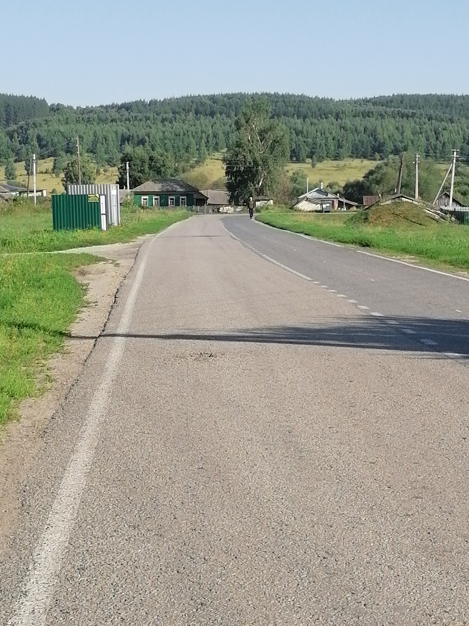 The image shows a paved road in a rural setting. On the left side of the road, there is a green corrugated metal fence bordering a grassy area. Beyond the fence, a green wooden house with white trim and several windows is visible. A single person, walking away from the camera, is on the left side of the road, near the fence. To the right of the road, there are fields and scattered buildings, including a small stone structure and a wooden shed. In the background, a densely forested hillside rises under a clear blue sky. Power lines and utility poles are present throughout the scene. The road has a white line marking the edge and a dashed white line separating lanes. The overall impression is a peaceful, sunlit rural landscape in or near the village of Bolshie Remezjonki, Russia.