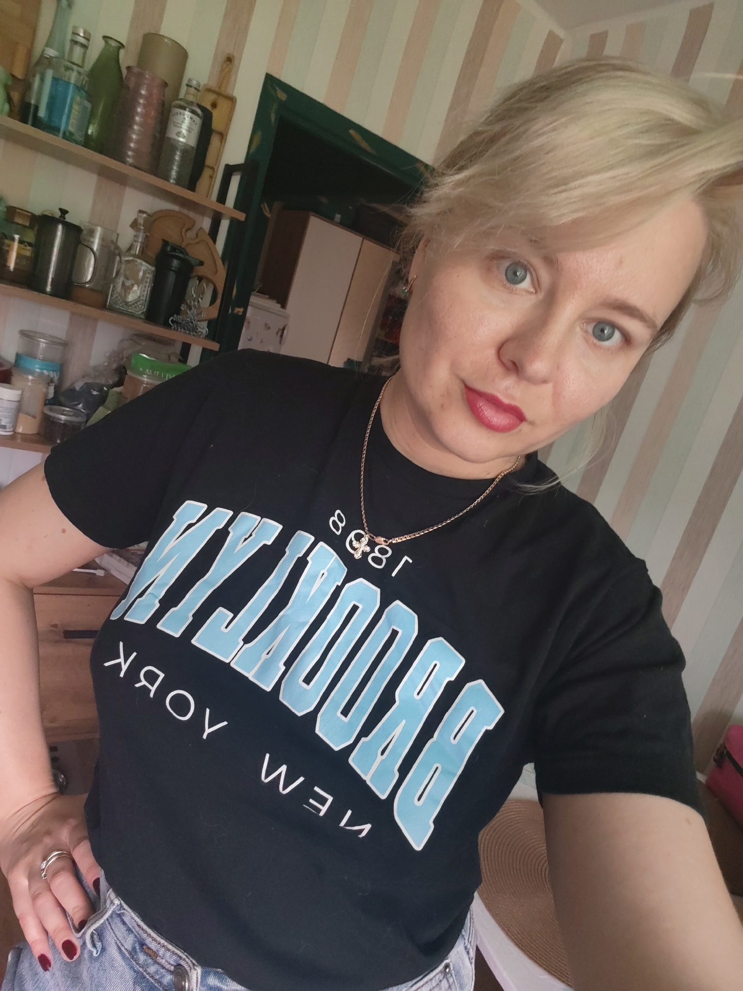 A blonde woman with blue eyes and red lipstick is taking a selfie. She is wearing a black t-shirt with the word "BROOKLYN" in light blue letters and "NEW YORK" below it, also in white letters. She is also wearing denim shorts and a gold necklace. The background appears to be an indoor setting with shelves of bottles and jars, suggesting a kitchen or pantry. The walls are striped with light and dark beige. The image quality is slightly grainy. There is no indication of the city of Izhevsk, Russia, or any other specific location.