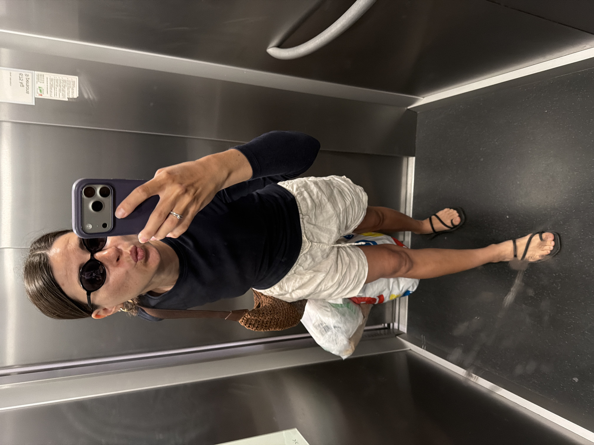 A person wearing sunglasses, a dark long-sleeved shirt, and white shorts takes a selfie in an elevator. They are holding a purple phone with multiple lenses. A ring is visible on their finger. They are carrying a straw bag and shopping bags. The elevator has stainless steel walls, a horizontal handrail, and a black floor. A sticker is affixed to the wall. The image appears to have been taken inside a building in Budva, Montenegro.