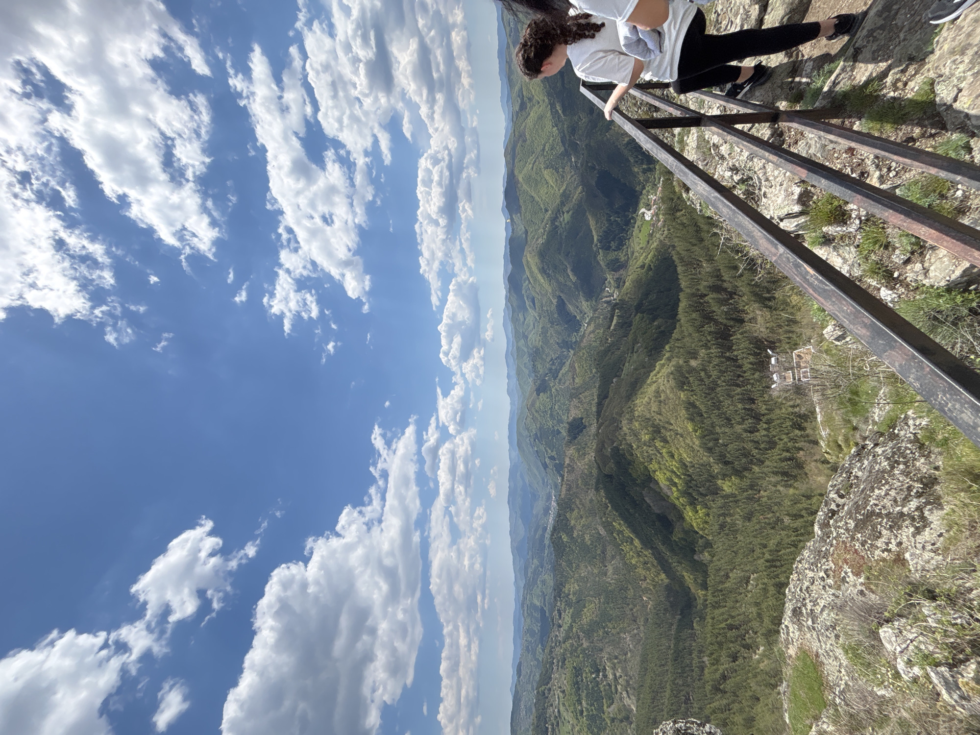 A high-angle, sideways view captures a scene from Gradat, Bulgaria. A person stands on a cliff edge, holding onto a rusted, metal railing. The landscape below consists of a vast, green valley with trees and a small village visible in the distance. The sky above is blue and filled with fluffy, white clouds.