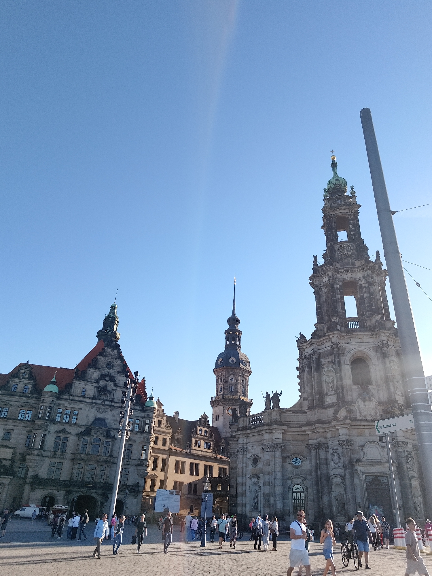 A low-angle shot captures a bustling public square in Dresden, Germany under a clear blue sky. Several notable buildings anchor the scene, including the Dresden Castle with its red-tiled roof and various towers, the Dresden Cathedral with its iconic Baroque tower, and other historical structures. The street sign “Altmarkt” is visible. The square is populated by numerous people of various ages, dressed in casual clothing. Many are walking across the square, suggesting a mix of locals and tourists exploring the area. A man is walking and pushing a bicycle to the right of the image. Sunlight illuminates the square and buildings, creating a bright and welcoming atmosphere.