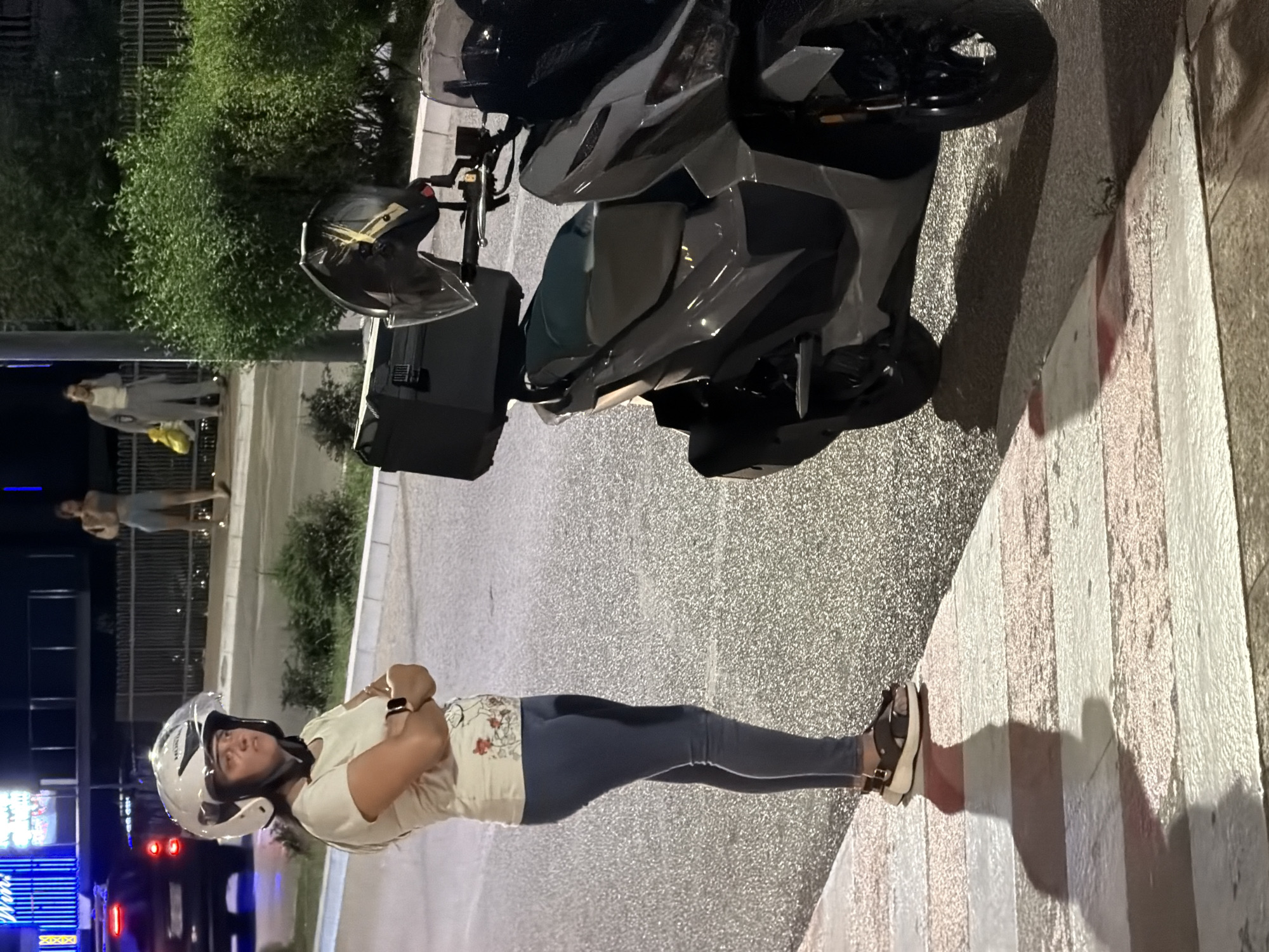 A woman wearing a white helmet and a light-colored t-shirt with floral designs, dark blue leggings, and brown sandals stands with her arms crossed on a crosswalk. She is looking to her left with a neutral expression. To her right is a dark gray scooter with a black storage box on the back. The scooter has a clear windshield. In the background, there is a dark-colored SUV with its taillights on, a metal fence, and lush green bushes. Beyond the fence, there are two individuals walking on a sidewalk. The scene appears to be at night, with streetlights illuminating the area. The location is identified as Budva, Montenegro.