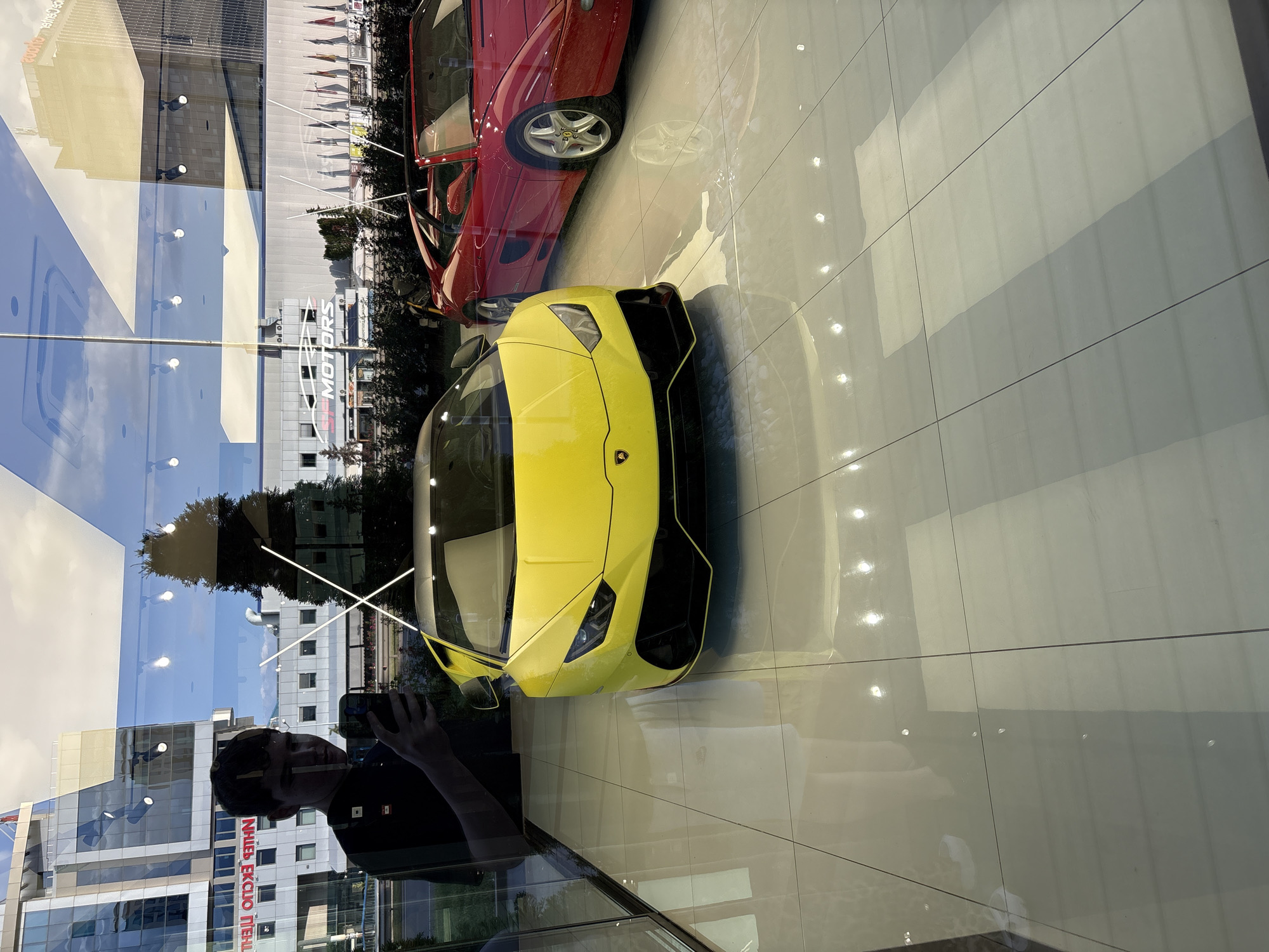 This image captures a vibrant yellow Lamborghini Huracan displayed inside a car showroom, viewed through a large, reflective glass window. Behind the yellow Lamborghini, part of a red sports car, possibly a Ferrari due to its classic lines and alloy wheel design, is also visible. Both cars are positioned on a glossy, light-tiled floor that reflects overhead lights and the vehicles themselves.
The glass window creates multiple layers of reflections, blending the interior scene with the exterior environment. A person is clearly reflected in the bottom left of the frame, holding a phone or camera to take the photograph, and wearing a dark t-shirt with what appears to be a small flag patch on the sleeve.
The reflections outside the showroom show a bright, clear daytime sky and various buildings, including one with prominent red text that reads "НПИБ ЕКСПО ПЛЕВЕН" (NPIB Expo Pleven), indicating a Bulgarian location. Lush green trees and other architectural elements are also reflected. Inside the showroom, a white sign with the word "MOTORS" is visible, further confirming the setting as a car dealership. The interplay of the bright yellow car, the sleek showroom, and the complex reflections from the outside creates a dynamic and visually engaging scene. The location is Sofia, Bulgaria.