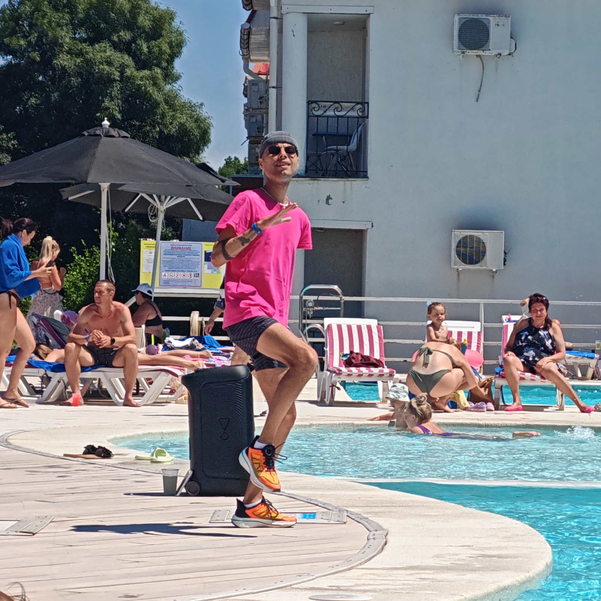 The image depicts a bright, sunny day at an outdoor swimming pool. A man in a pink t-shirt, grey beanie, and sunglasses is posing on the wooden deck, with his right leg bent and his left arm extended. He is wearing striped shorts and bright orange sneakers. Next to him is a large black portable speaker.
Several other people are visible around the pool. To the left, a woman in a blue bikini top and black bottoms is holding a phone. Behind her, a woman with blonde hair is also looking at her phone. Further back, a man in dark shorts and no shirt sits on a lounge chair, holding his hands together. Another man is partially visible in the background, also on a lounge chair.
In the pool, a child is seated on the edge, and a woman in a green bikini is crouched near them. Further into the pool, a woman with her hair in a bun swims. Other lounge chairs with people are visible in the background, some with red and white striped cushions. A sign with text in what appears to be Cyrillic script is mounted on a pole. The building in the background is white with balconies and an air conditioning unit. The ground is a light-colored wooden deck, with a curved edge leading to the blue pool water.