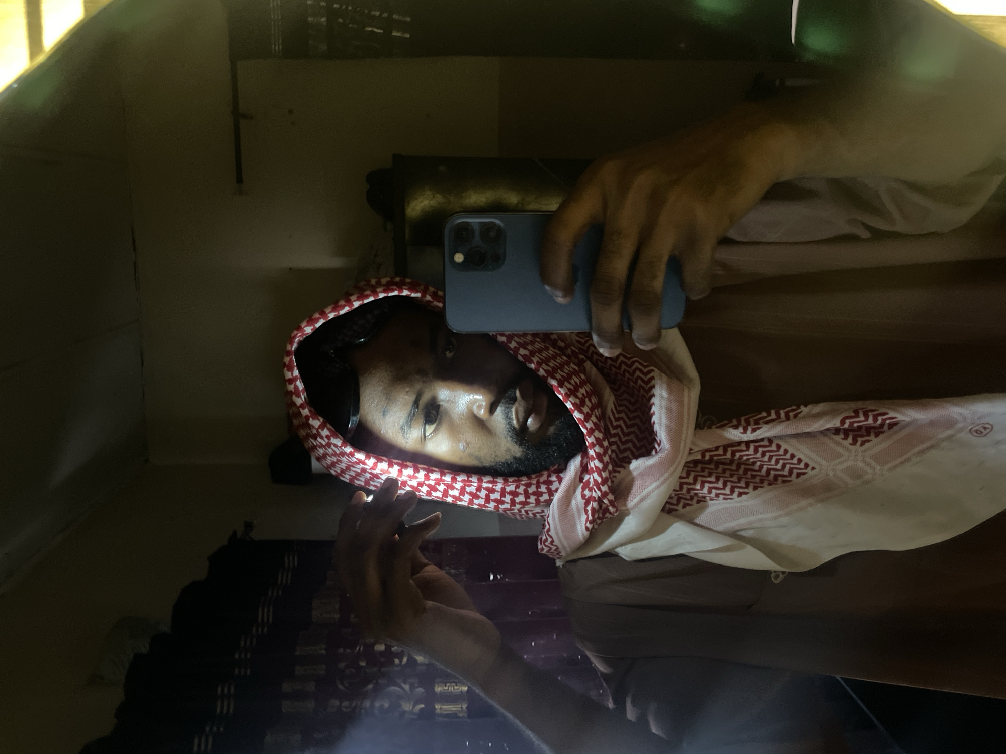 A man with dark skin, a beard, and wearing a red and white patterned headscarf is taking a selfie in a mirror with a blue phone. The reflection shows a cluttered interior space, potentially a small room, with various items on shelves and possibly a curtain or blinds in the background. It is not possible to identify the location as Lokoja, Nigeria, or any other specific place based on the visual information provided.