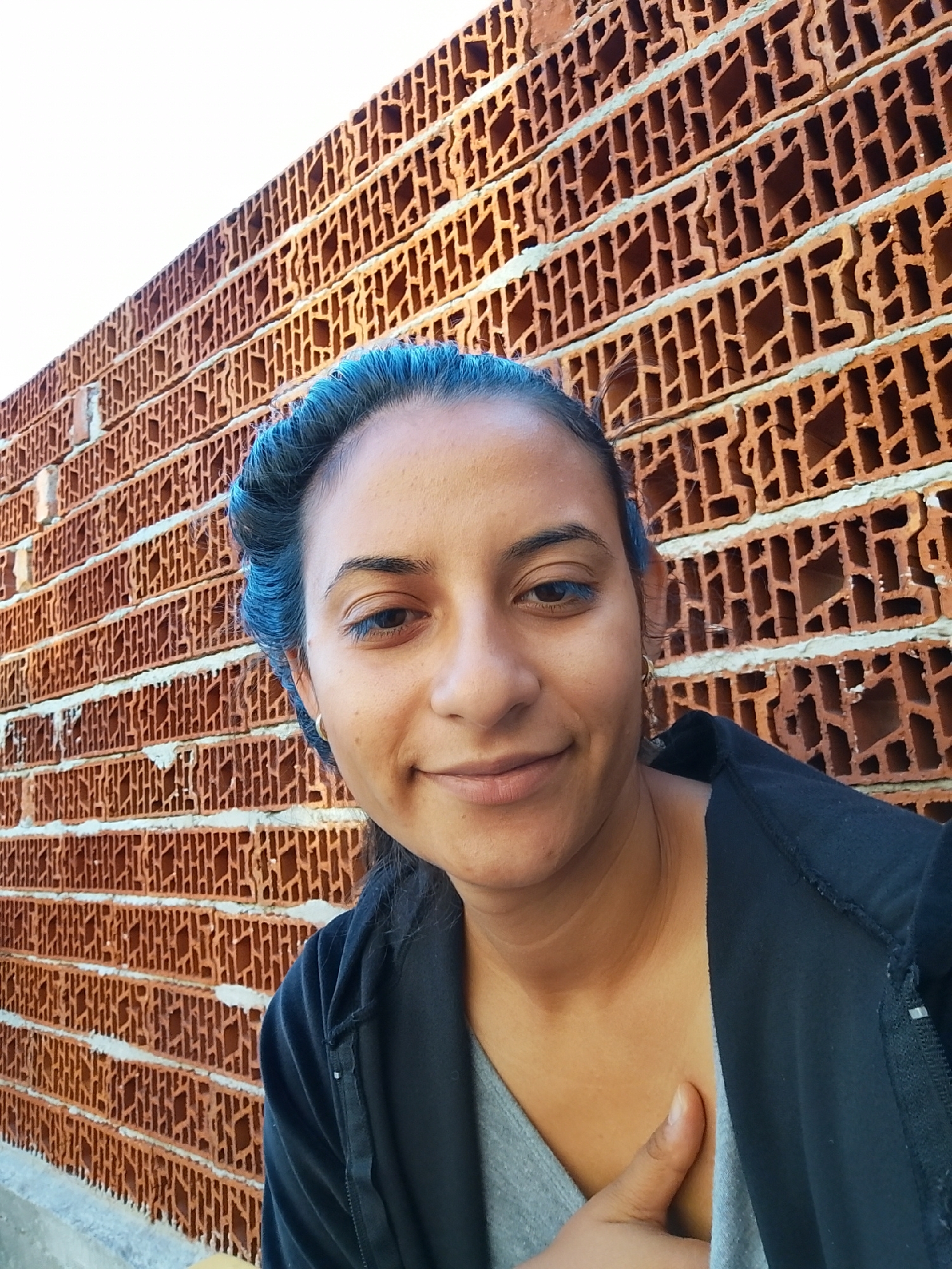 A selfie of a woman with dark hair styled up, featuring blue eyeliner, is taken against a background of a brick wall. The woman is wearing a black zip-up jacket over a grey top, and her right hand is placed on her chest. The brick wall is made of hollow, orange-brown bricks stacked horizontally, with white mortar visible between them. The scene appears to be outdoors, with a bright, white sky visible in the upper left corner. The location is identified as Yambol, Bulgaria.