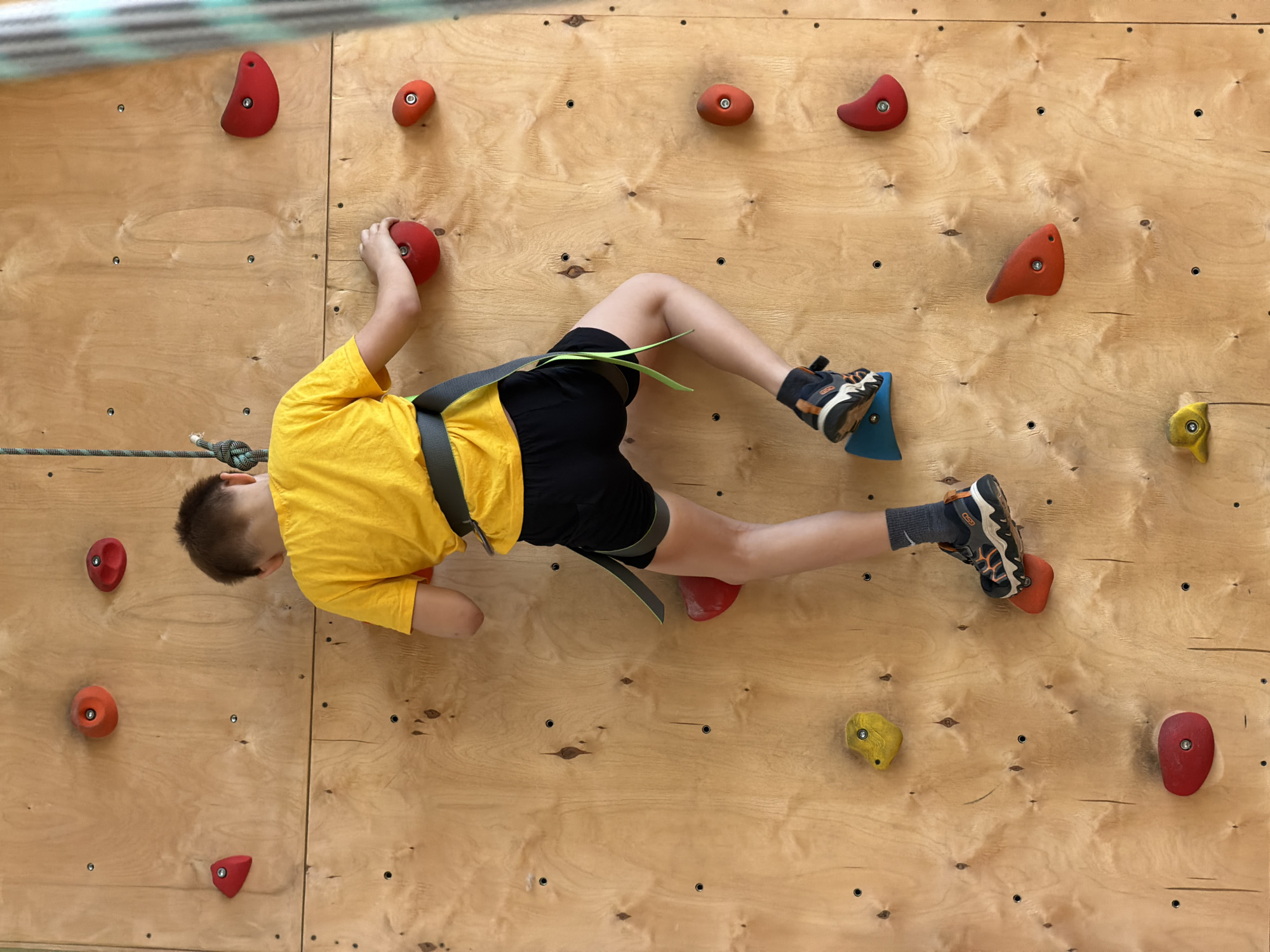 A child is shown climbing on a wooden climbing wall indoors. The child, wearing a yellow shirt, black shorts, gray socks, and athletic shoes, is secured with a climbing harness and rope. The climbing wall is fitted with multiple red, orange, and yellow climbing holds. The location may be in the city of Izhevsk, Russia, though no details in the image confirm it.