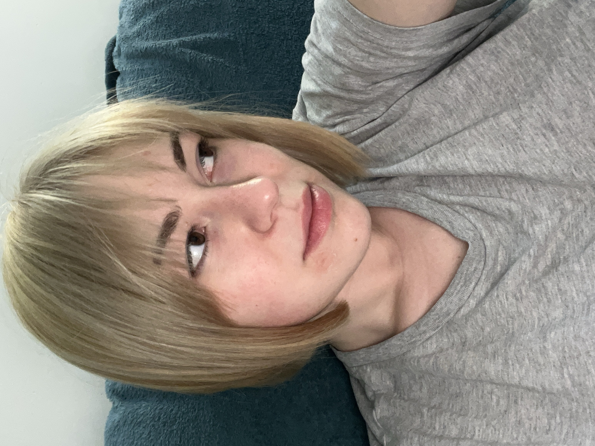A young woman with blonde hair is lying down on a blue surface, possibly a pillow or blanket. She is wearing a grey t-shirt. Her eyes are brown and she is looking upwards. The background appears to be a white wall. The location in the city of Boreti, Montenegro is not discernible from the image.