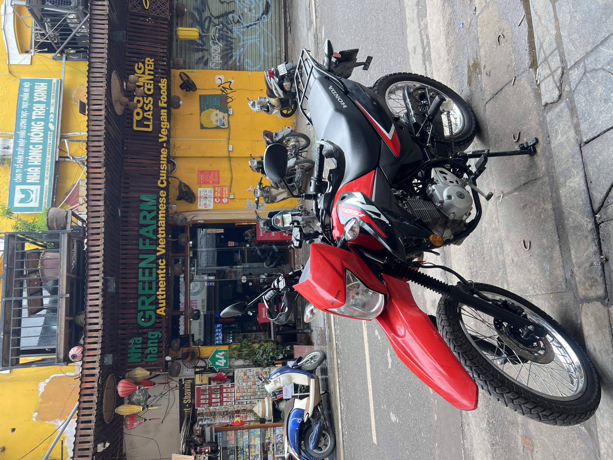 The image captures a street scene in Hanoi, Vietnam, likely in a commercial district. In the foreground, a red and black Honda motorcycle is parked on the sidewalk, its front wheel and fender prominently displayed. The motorcycle has a license plate that is partially visible. Behind the motorcycle, a blue and white scooter is parked.
The background reveals a yellow building with a prominent sign that reads "NHÀ HÀNG GREEN FARM AUTHENTIC VIETNAMESE CUISINE - VEGAN FOODS" in green and yellow lettering. Another sign above it, in blue, also advertises the business. Several paper lanterns in red and pink hues hang from the building's overhang. Further back, various shops and storefronts are visible, with some displaying goods on shelves. Graffiti adorns the wall of one of the buildings. Several other motorcycles are parked along the street, and a few individuals are discernible within the shops. The street itself appears to be paved, with some wet patches on the sidewalk, suggesting recent rain. The overall atmosphere is that of a busy urban environment with a strong presence of local businesses and daily life.