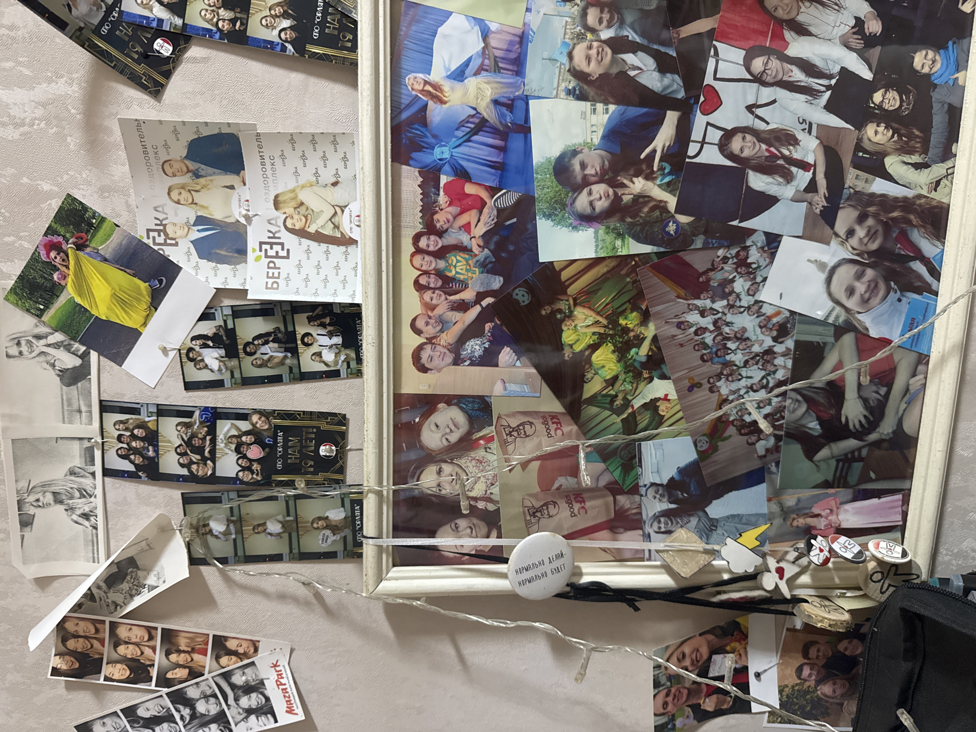 The image displays a vibrant collage of numerous photographs and memorabilia arranged on a light-colored, patterned wall, suggesting a personal display board or a section of a room. The central feature is a large white picture frame, also filled with a dense collection of photos. String lights with small bulbs are draped horizontally across the lower part of the frame and extend to other areas of the wall, adding a decorative touch.
The primary subjects in the photos are young people, predominantly teenagers and young adults, often smiling, posing, and interacting in groups. Their expressions generally convey joy, camaraderie, and happiness. The setting varies across the photos, including what appears to be outdoor natural environments, indoor event spaces, and possibly institutional or camp settings.
Various objects are integrated into the collage:
* Numerous photo prints of different sizes, including standard prints and several photo booth strips (some in color, some black and white).
* Small decorative elements like a white circular pin, a yellow lightning bolt, a white cloud, a red heart, a star, and small wooden discs.
* A black pouch or bag is visible at the bottom right.
* One photo shows individuals holding KFC "sogood" paper bags.
The photos depict a range of activities, suggesting a collection of memories from social gatherings, camps, celebrations, and outings. There's a strong sense of shared experiences and friendships.
Visible text includes:
* "Maza Park" on a photo booth strip.
* Repeated instances of "СПО 'ОРЛЯТА'" (SPO 'Orlyata'), likely referring to a youth organization or camp.
* "НОРМАЛЬНО ДЕЛАЙ - НОРМАЛЬНО БУДЕТ" (Do it normally - it will be normal / Do good - it will be good) on the white circular pin.
* "KFC sogood" on paper bags in some photos.
* "БЕРЁЗКА" (Beryozka, possibly a camp or complex name) along with "ОЗДОРОВИТЕЛЬНЫЙ КОМПЛЕКС" (Health Complex / Wellness Complex) appears on several promotional-style prints.
* "НАМ 19 ЛЕТ!" (We are 19 years old!) is visible on multiple photo strips and prints, possibly indicating a celebration or anniversary.