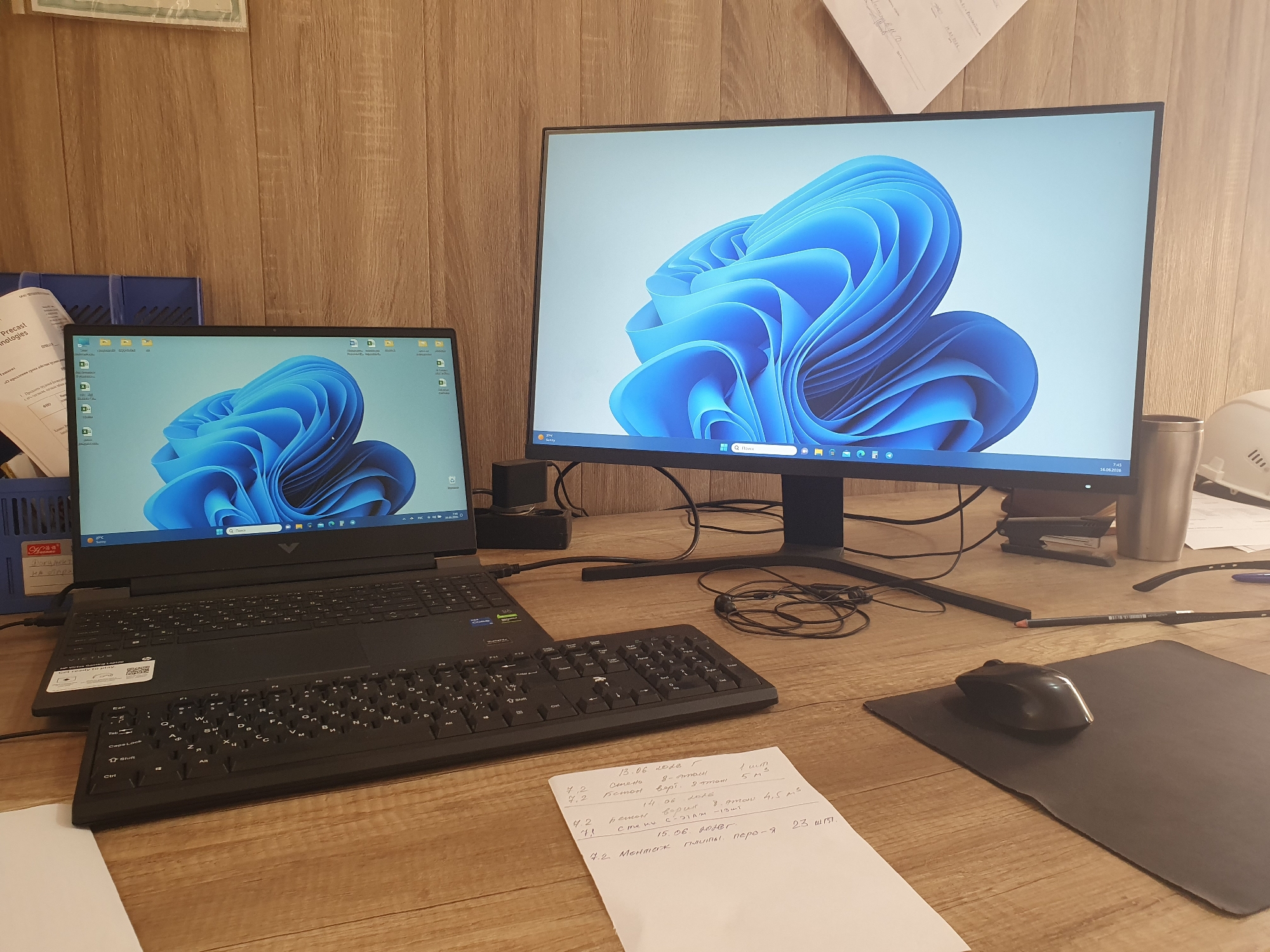 The image shows a desk with two computer monitors and a laptop, all displaying the same Windows 11 wallpaper. A separate keyboard is placed in front of the laptop, and a mouse rests on a black mousepad to the right. A crumpled piece of paper with handwritten notes is on the desk. In the background, a wooden paneled wall is visible, with some papers and office supplies scattered around. A white hard hat is also present on the right side of the desk. The scene appears to be an office or workspace. There is no indication of the location being Tashkent, Uzbekistan.
