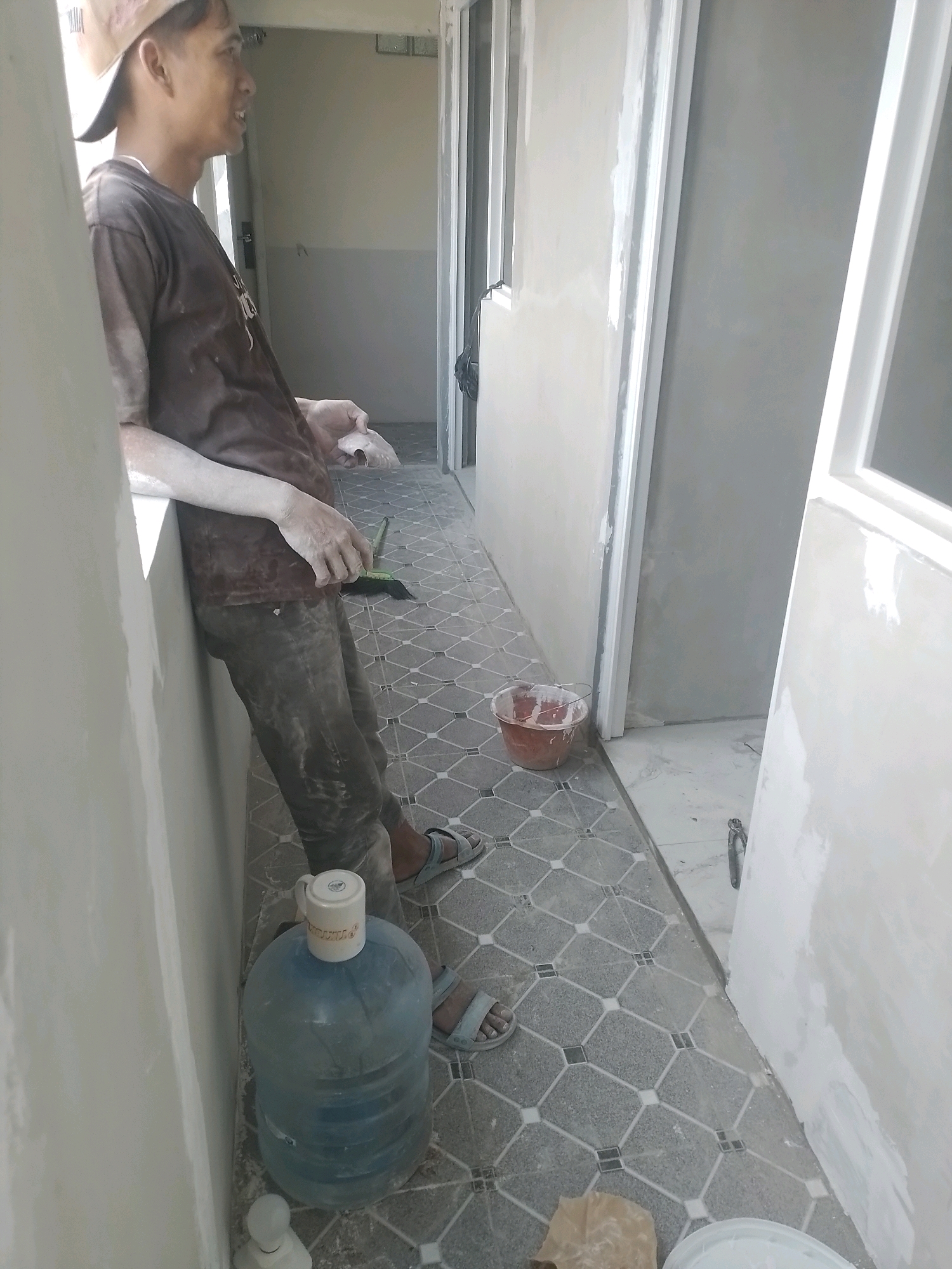 A man wearing a brown t-shirt and a beige baseball cap leans against a light-colored wall. His arms and clothes are dusted with white powder, suggesting he is engaged in construction or renovation work. He is holding a white object, possibly a piece of plaster or putty, in his right hand. A broom with a green handle lies on the floor near his feet. The floor is tiled with a geometric pattern of gray and off-white squares. In the background, a red bucket sits on the floor, and a large blue water dispenser with a white mug on top is positioned near the man. Two doorways are visible, leading into dimly lit rooms. The environment appears to be an interior hallway or unfinished room, consistent with a construction site in West Jakarta, Indonesia.