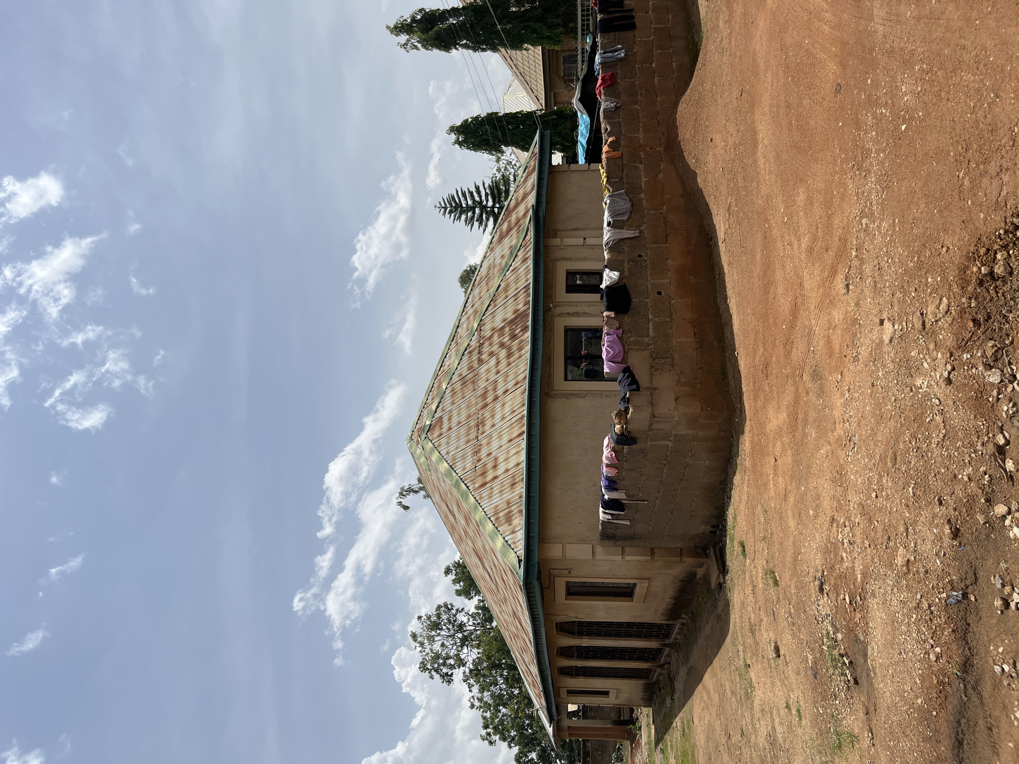 In the city of Bukuru, Nigeria, a modest, single-story house stands under a partly cloudy sky. The house features a weathered, corrugated metal roof with a green trim. The walls are made of a light-colored, textured material, and several arched windows with dark bars are visible on the side facing the viewer. In front of the house, a low wall constructed from concrete blocks serves as a clothesline, displaying a variety of drying laundry, including shirts and pants in different colors. The ground in front of the house is a reddish-brown dirt, scattered with small stones and debris. Trees and other structures are visible in the background.