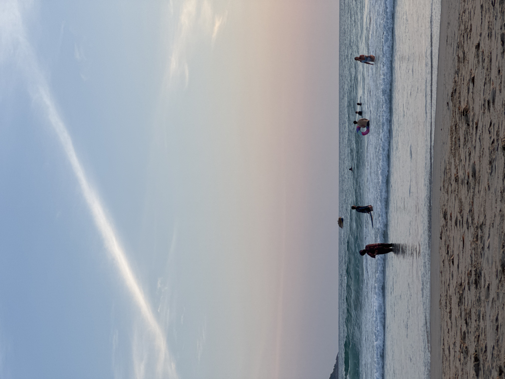 The image depicts a serene beach scene at Karon Beach, Thailand, likely during dusk. The sky is a gradient of soft blues and pinks, with a wispy cloud formation cutting across the upper portion. The sea is calm with gentle waves rolling onto the sandy shore. In the water, several individuals are visible. One person stands in the shallows on the left side of the frame, facing away from the camera. Further out, a few more people are wading or swimming, one of whom has a colorful inflatable ring. In the distance, a small boat is visible on the horizon. The foreground consists of the sandy beach with subtle textures of shells and debris. The overall atmosphere is peaceful and tranquil.