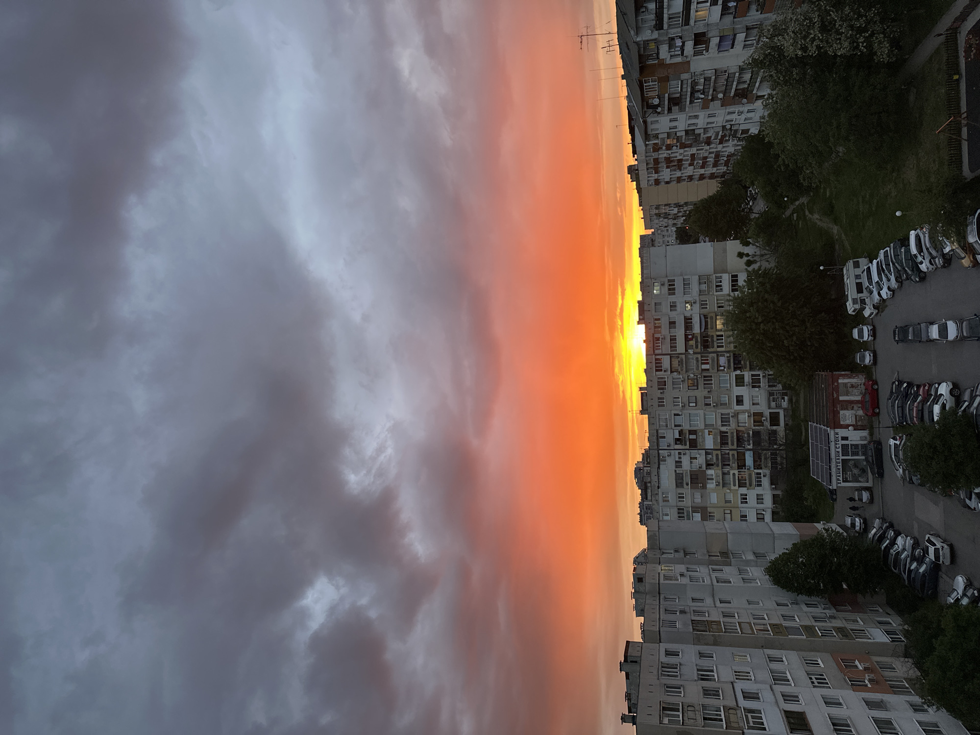 The image shows a sunset view from an elevated position, possibly a high-rise building, overlooking a residential area in Sofia, Bulgaria. The sky displays a dramatic contrast between dark, overcast clouds on the left and a vibrant orange and yellow horizon line where the sun is setting or has recently set. To the right, dense, Soviet-era apartment blocks dominate the landscape. A parking lot is visible with numerous parked vehicles, primarily white and gray in color, alongside the buildings. Trees are interspersed among the buildings, adding patches of green to the urban setting. Electrical lines and antennas can be seen on the rooftops of some buildings. The scene is generally still and quiet, reflecting the moment before nightfall.