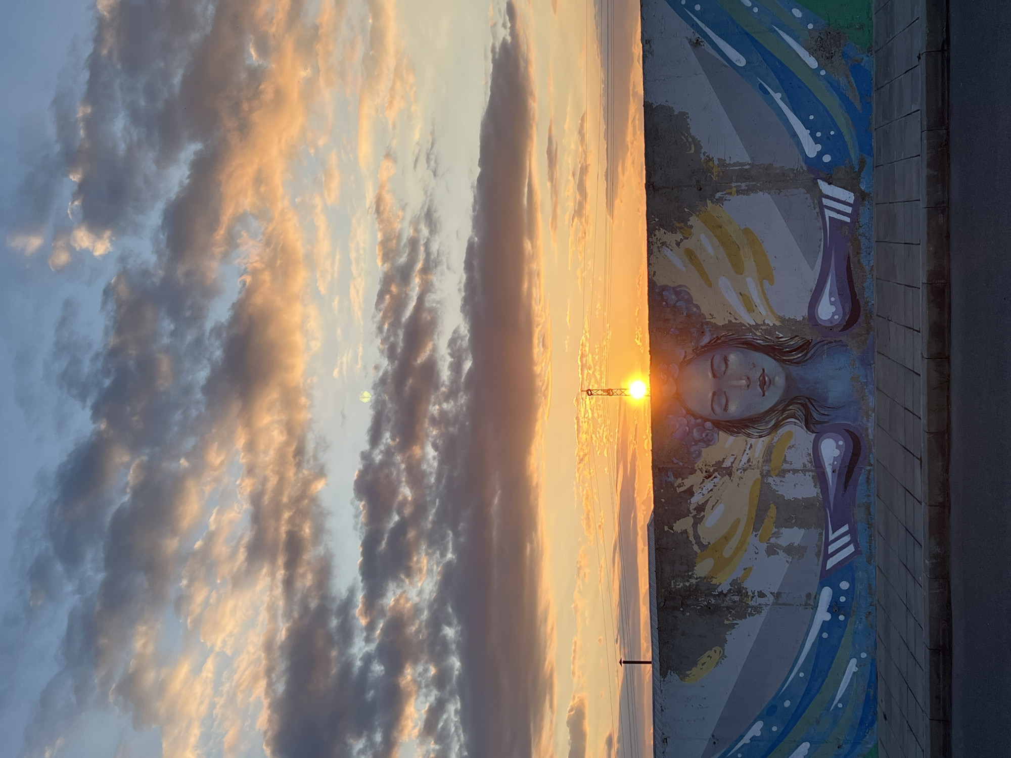 A vertical eye-level shot shows a sunset in Burgas, Bulgaria. The sky is filled with clouds, some darkened and others illuminated by the setting sun with orange and golden hues. In the foreground, there is a concrete wall featuring a mural of a woman with closed eyes and long, flowing hair. The mural includes elements of blue and yellow, suggesting wings. The sun sits directly in front of the mural, casting a glare. Power lines cross the sky. A dark, thin pole is just below the sun. The edge of a sidewalk is visible on the right.