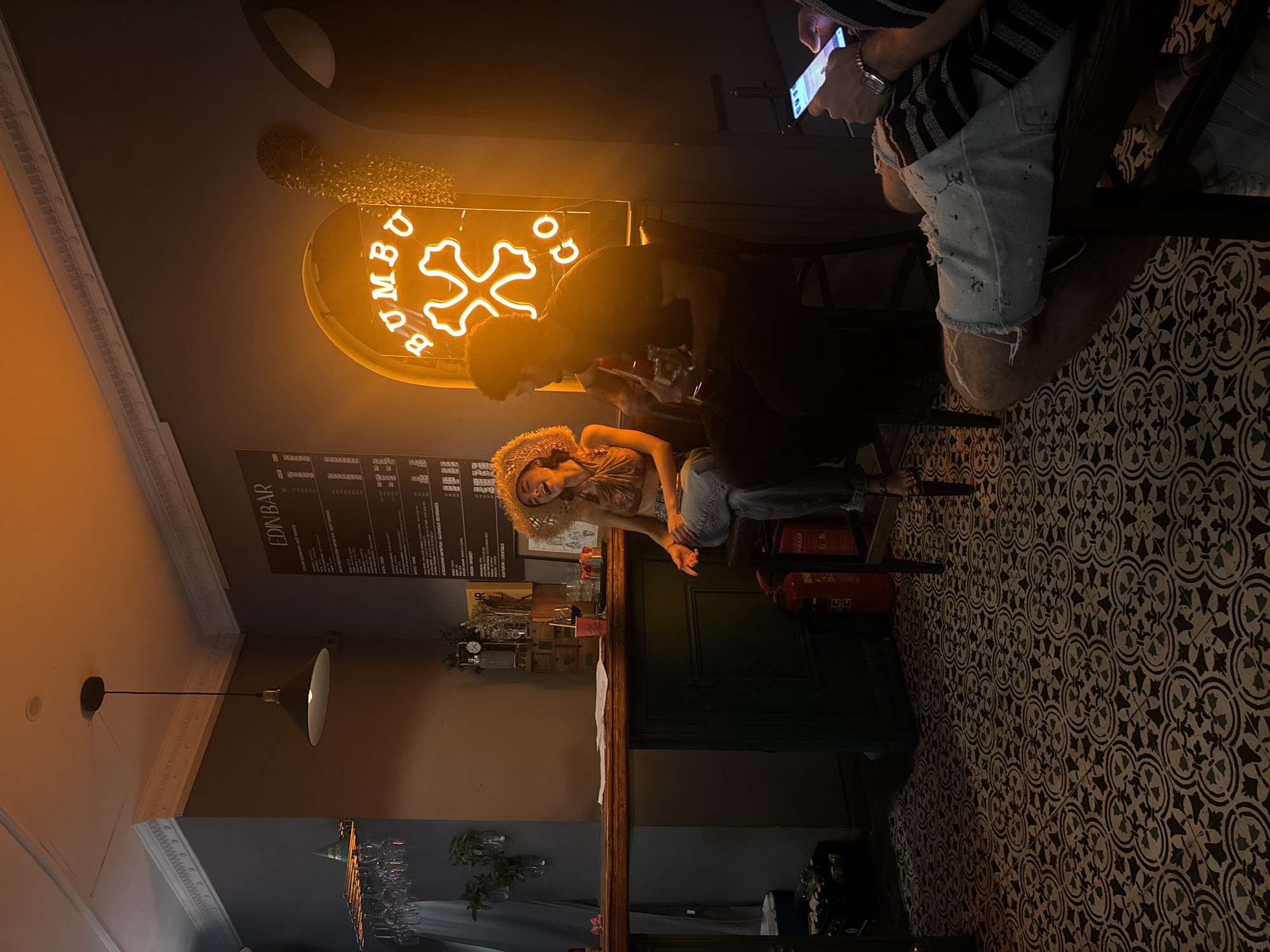 The image depicts an interior scene at "EDIN BAR" in Sofia, Bulgaria. The bar features a prominent neon sign that spells "BUMBU GO" in an orange glow. Two individuals are seated on stools at the bar. One, a woman with curly hair and wearing a straw hat, is actively engaged in conversation or laughter, leaning back on her stool. She is dressed in a brown halter top and blue jeans. The second person, a man, is partially obscured and appears to be holding a bottle. A third person, seated further to the right and out of focus, is looking at their phone. The bar itself is made of wood, with a green counter base. The floor is decorated with an intricate black and white tiled pattern. The overall ambiance is dimly lit, with the neon sign providing a strong source of light. Other visible objects include glassware on a rack above the bar, a fire extinguisher, and a menu board on the wall.