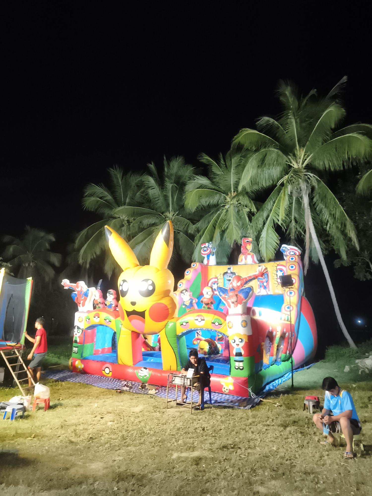 In Baan Bo Phut, Thailand, at night, a large, colorful inflatable castle featuring characters like Pikachu and superheroes is set up on a grassy area. Several people are present. To the left, a young person in a red shirt stands on a ladder leading to an entrance of the inflatable. In the center, a young person in black sits at a small table in front of the inflatable. To the right, a young person in a blue shirt and dark shorts sits on a small stool, looking at their phone. The background shows several palm trees against a dark sky. The ground is covered in grass, and a blue and white striped tarp is spread out beneath the inflatable. Several small stools and bags are scattered around the area.