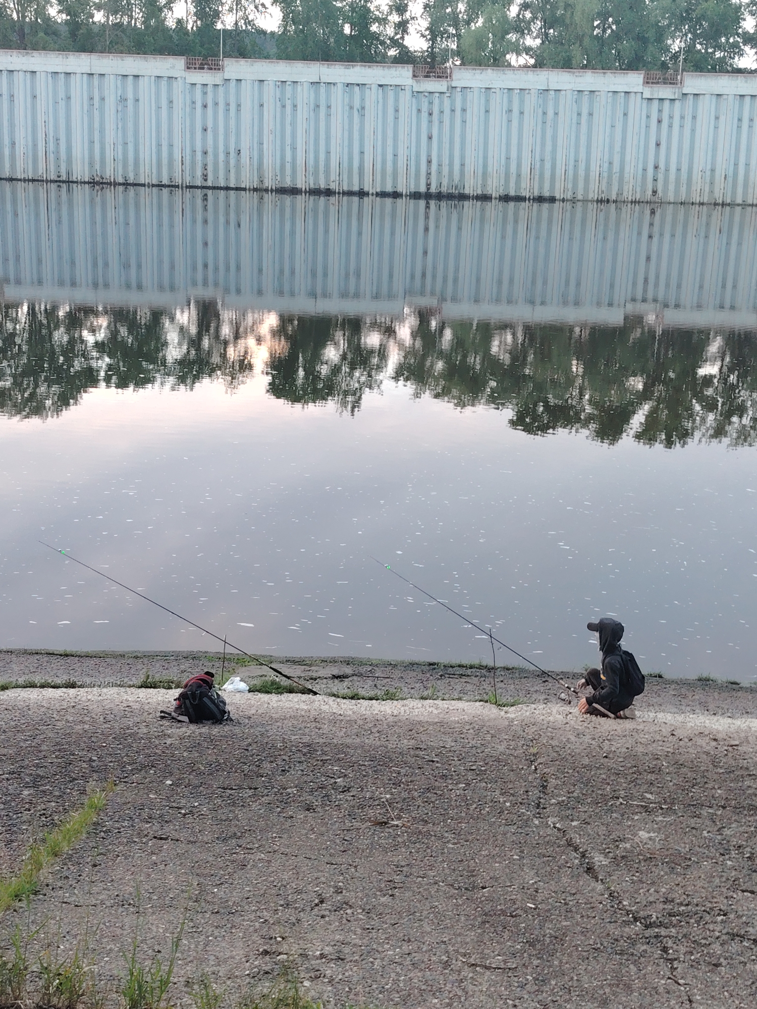 The image depicts an outdoor scene of two individuals fishing by a calm body of water, likely a river or canal. In the foreground, a concrete bank slopes down to the water's edge where the two people are positioned. The person on the left is mostly obscured by a dark backpack and a white bag, with a fishing rod extending into the water. The second individual, on the right, is crouched, wearing a dark hooded top and a cap, holding a fishing rod that also extends into the water.
Across the water, a long, light-colored metal wall, resembling sheet piling, forms the opposite bank. This wall reflects distinctly on the water's surface, creating a mirror image. Above the wall, a dense line of green trees is visible, with their foliage also reflected in the water. The water itself is smooth, with minor ripples and small scattered white specks.
The lighting suggests either dawn or dusk, as a soft, warm glow, possibly from the sun low in the sky, is reflected on the water, particularly noticeable in the center. The sky is partially visible above the trees, appearing bright. The scene conveys a sense of peaceful activity, with the individuals focused on fishing. No discernible text is visible in the image. The setting combines natural elements with man-made structures along the waterfront in Chaikovsky, Russia.