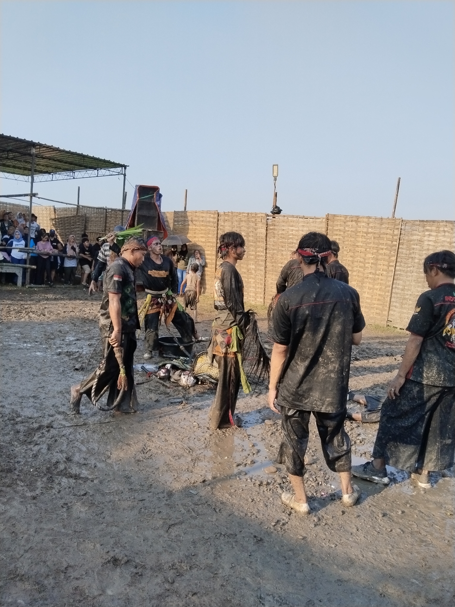 A group of men, splattered with mud, stand in a muddy field in Kedawung, Indonesia. They are wearing dark shirts and pants, some with red headbands. In the background, a crowd watches from under a shelter and behind a woven bamboo fence. Miscellaneous objects are scattered on the ground near the men.