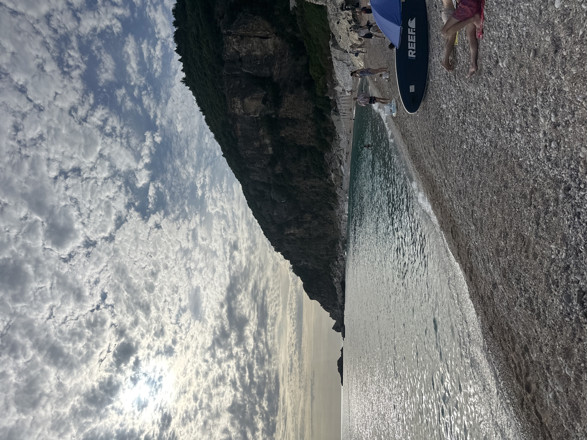 The image depicts a coastal scene at Mirište, Montenegro, under a sky largely covered by patchy altocumulus clouds. The sun is partially obscured but visible on the upper left, casting a bright glare and creating reflections on the water.
The left side of the frame features a calm, reflective sea with a smooth surface broken by minimal ripples and small, gentle waves breaking on the shore. The water appears dark grey with streaks of bright light reflecting the sky. In the mid-distance on the left, several small, dark rock formations or islets are visible in the sea, along with a faint line of buoys extending horizontally.
The right side of the image is dominated by a steep, dark cliff face or mountain, heavily vegetated with trees and shrubs on its upper slopes. The lower sections of the cliff reveal exposed rock formations, dark grey and brown in color, meeting the beach directly.
The foreground and mid-ground consist of a pebble beach, composed of numerous small to medium-sized grey and white stones. The beach extends from the left into the mid-ground and forms the base of the cliff on the right.
Multiple individuals are present on the beach and in the water. On the far right foreground, two pairs of human legs are visible, belonging to individuals lying on red towels on the pebbles. Adjacent to them, a dark blue stand-up paddleboard (SUP) with "REEF" branding in white text is positioned on the beach. A blue beach umbrella is partially visible next to the SUP.
Further along the shoreline towards the center-right, several people are standing on the pebbles or at the water's edge. One individual is partially submerged in the water. A person holds a small child near the waterline. A white inflatable ring lies on the pebbles. Additional figures are dispersed along the beach towards the distant cliff base, engaged in various leisure activities such as walking and observing the surroundings.