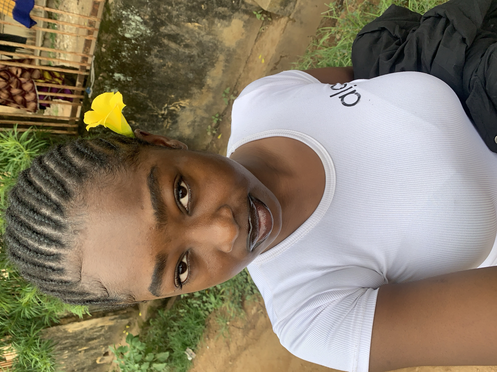 A medium shot captures a young Black woman from the chest up, looking directly at the camera. She has braided hair with small baby hairs along her hairline and a bright yellow flower tucked behind her right ear. She is wearing a white, ribbed t-shirt with a black logo on the left side of her chest, which appears to read "alo". Her lips are glossy and dark. To her right, a portion of a black jacket or garment is visible, draped over her shoulder. The background consists of an outdoor scene with a textured, light-colored wall on the left, lush green foliage, and a dirt path. In the background, a wooden fence with vertical slats is visible, with colorful clothing hanging behind it, suggesting a residential or market area. The overall impression is a candid portrait taken outdoors. The presence of the fence and the style of clothing in the background suggest a location in Nigeria, possibly in the city of Rafin Par, Nigeria.