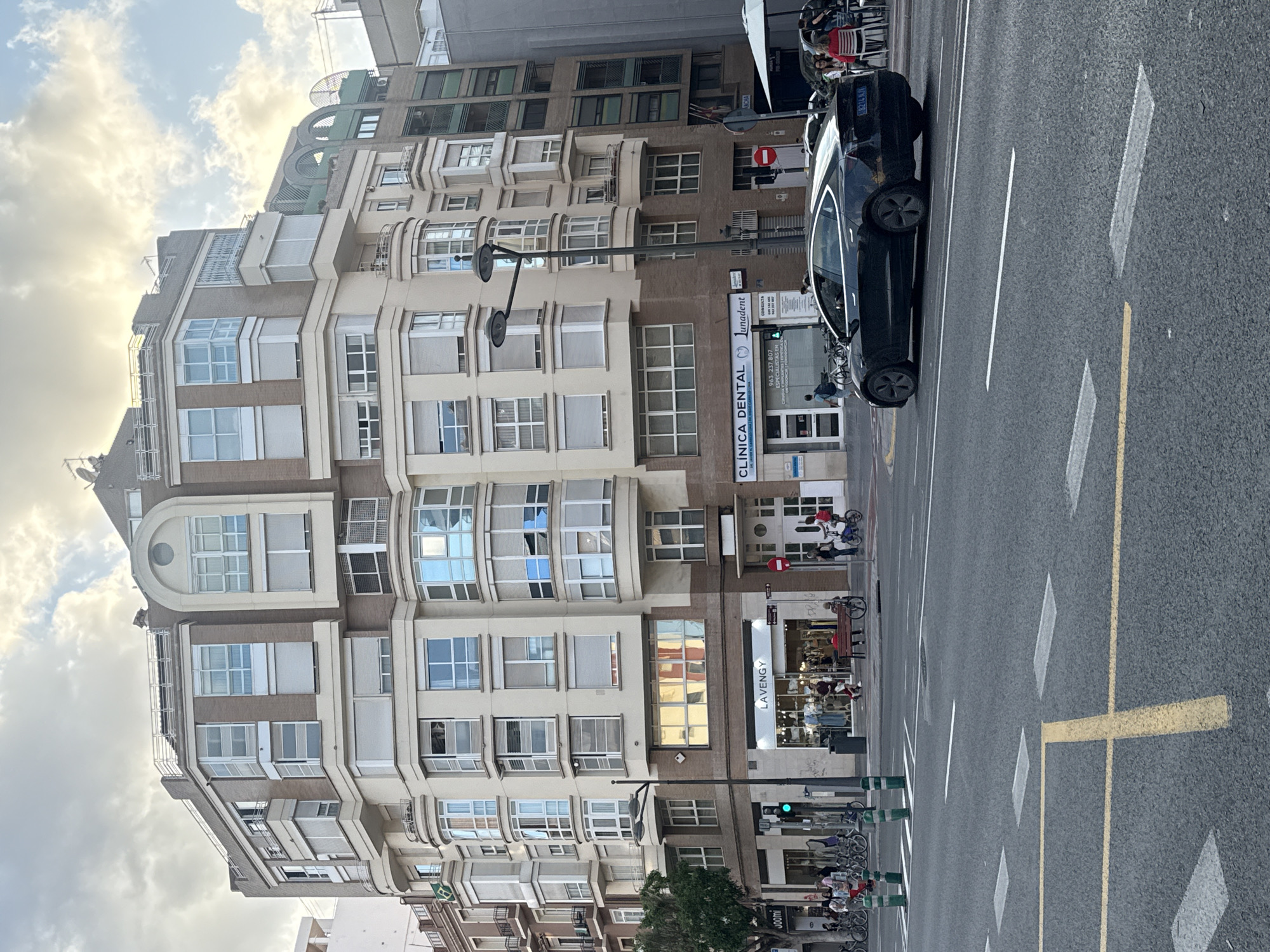 This is a street-level, eye-level, medium-distance, full-body shot of a street scene in Valencia, Spain. A black Tesla Model 3 sedan drives away from the camera on the right side of the road. The car is in the middle lane of a three-lane road, with yellow and white lane markings visible. The building in the background is a multi-story commercial and residential building with a light beige facade and dark brown brick accents. The building features a rounded corner with multiple bay windows. Several shops are visible at street level, including a clothing store labeled "LAVENGY" and a dental clinic labeled "CLINICA DENTAL" with the text "Lunadent" also visible. Streetlights and traffic signals are present. Several bicycles are parked on the sidewalk in front of the shops. In the foreground, the road surface is asphalt with pedestrian crosswalk markings. The sky is cloudy with some patches of sunlight.