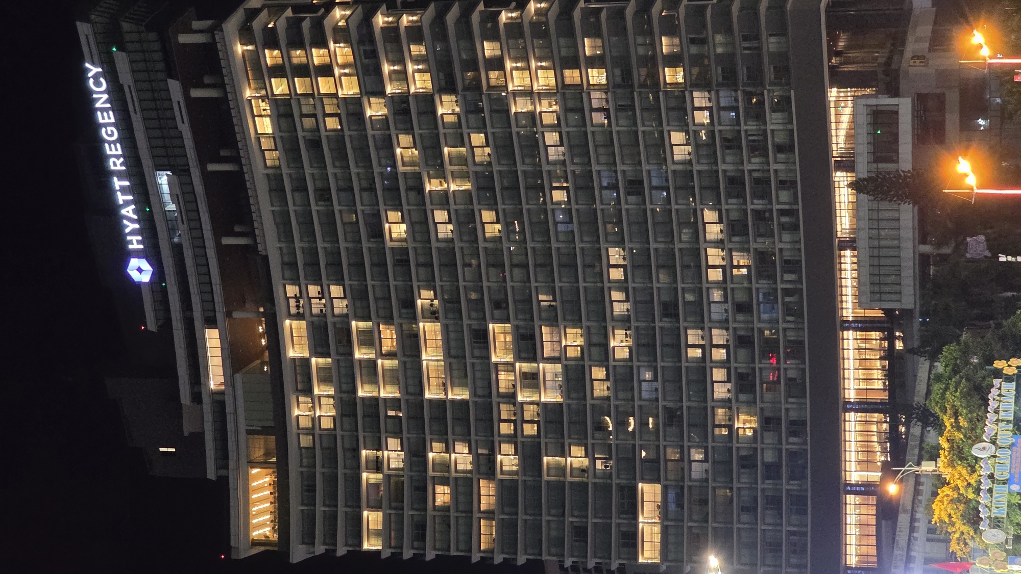This image captures a large, modern building at night, likely a hotel, in Nha Trang, Vietnam. The building dominates the left and central parts of the frame, stretching vertically with numerous windows. The dark sky indicates it is nighttime, and many of the building's windows are illuminated with warm, yellow light, creating a striking contrast with the darker exterior.
A notable detail is the pattern formed by the illuminated windows on the building's facade. Specifically, in the central section, a heart shape is visible above the letters "VN," suggesting a patriotic or welcoming message for Vietnam.
On the left side of the building, a large, illuminated sign vertically spells out "HYATT REGENCY" with the distinctive Hyatt logo. To the right of the main building, at ground level, there are additional structures and bright outdoor lighting, including streetlights with intense orange glows and decorative lights on trees. Another illuminated sign at street level displays the Vietnamese text "KÍNH CHÀO QUÝ KHÁCH," which translates to "WELCOME, DEAR GUEST." The overall scene portrays a vibrant urban environment after dark.