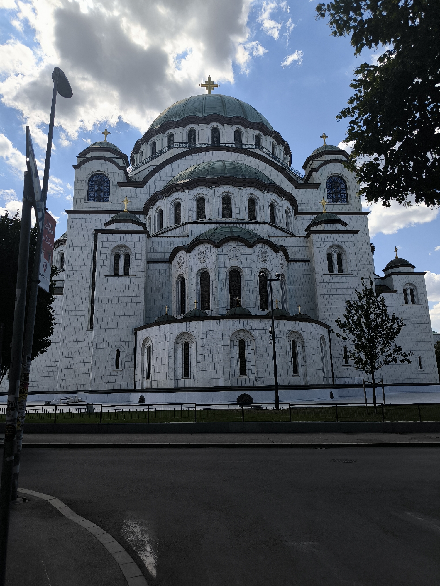 The image shows the exterior of the Saint Sava Temple, a prominent Orthodox church in Belgrade, Serbia, viewed from a street. The massive white stone building features multiple domes topped with golden crosses, arched windows, and intricate architectural details. The facade is clad in white marble blocks, giving it a bright and imposing appearance. A dark green railing runs along the front of the church, separating it from a patch of manicured lawn. A tree stands to the right of the church, its branches extending into the frame. In the foreground, a dark asphalt road curves to the left, with a concrete curb and sidewalk. A tall lamp post and a signpost are visible on the left side of the image, partially obscuring the view of the church. The sky is blue with scattered clouds, and the sunlight suggests it is daytime. The scene is devoid of people.