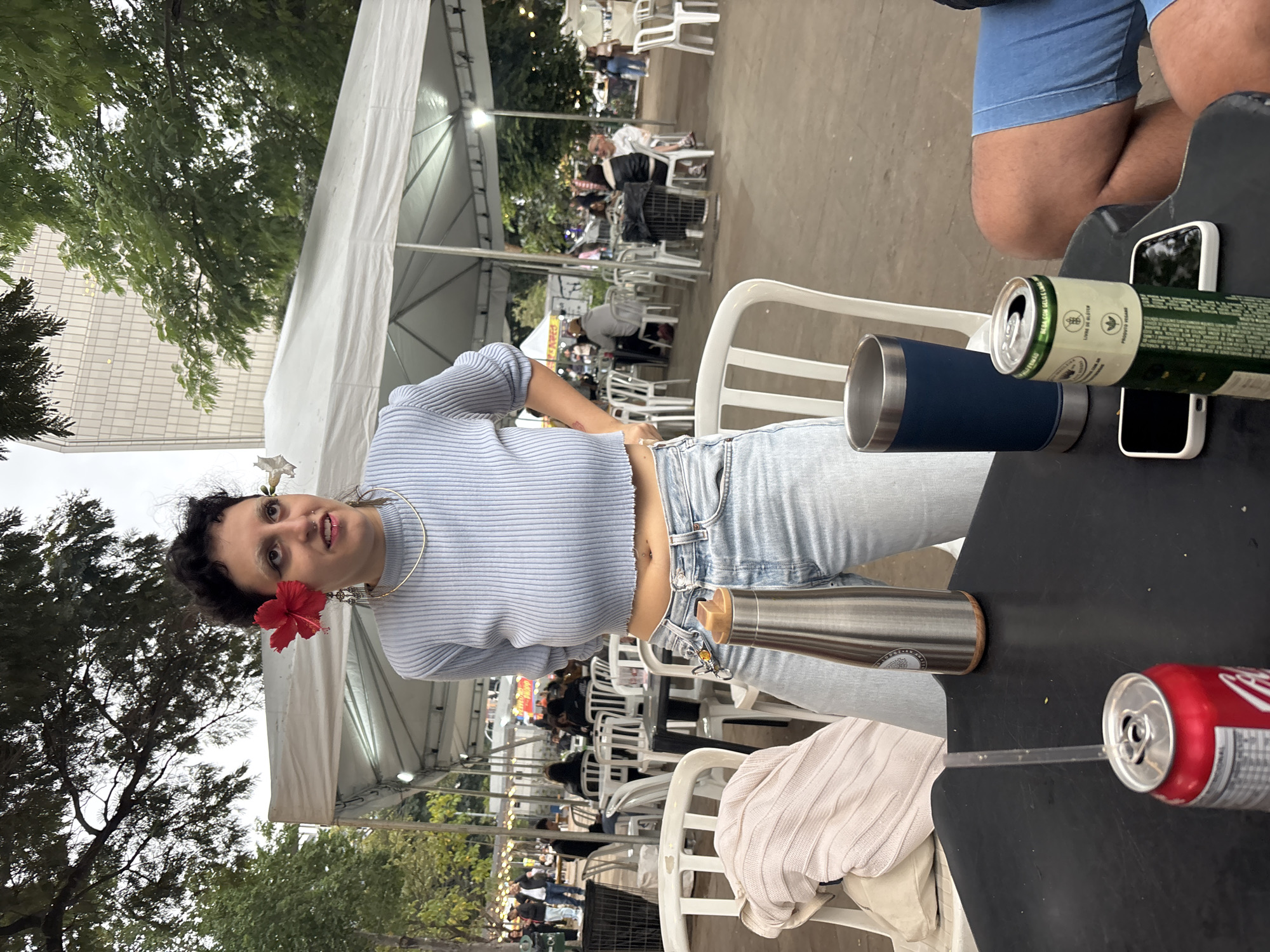 The image shows a woman at an outdoor event, possibly a fair or market, in what appears to be São Paulo, Brazil. The setting is underneath a white tent structure with visible support beams. The background is filled with people sitting at white chairs and tables.
The woman is wearing a light blue, long-sleeved, striped shirt, light blue jeans, and has two flowers in her hair, one red behind her ear and one white above it. She also has a gold choker necklace.
On the table in front of her, there are two metal water bottles, an open can of what looks like a Coca-Cola with a straw, and an open can of beer. There is also a cell phone. Another person wearing blue shorts is partially visible at the top right corner of the image.
