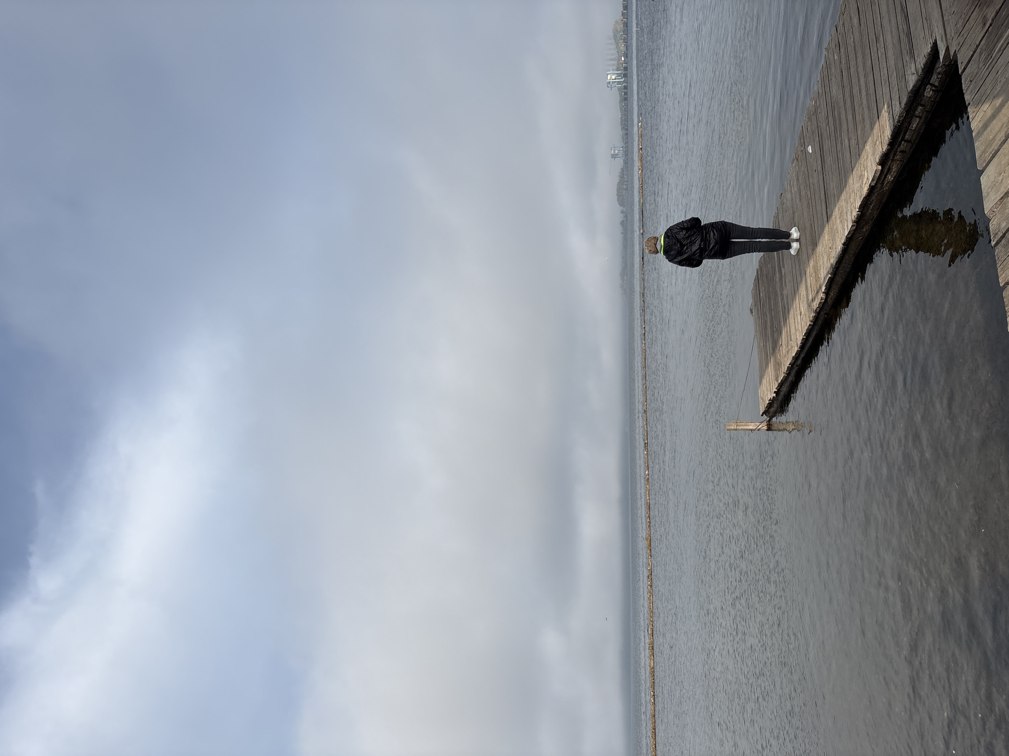 A person stands on a wooden dock, facing away from the camera, looking out at a body of water. The person is wearing a black hooded jacket with a green collar, dark pants, and white shoes. The sky is overcast with clouds. In the distance, there is a hazy horizon with some structures visible, possibly industrial buildings. The water is calm, with a reflection of the dock and the person visible on its surface. The dock is made of weathered wooden planks. The scene suggests a cool or foggy day, with a sense of solitude and contemplation. The specific location of Ust-Ilimsk, Russia, is not directly identifiable from visual cues within the image itself, as it is a general outdoor scene without distinguishing landmarks.