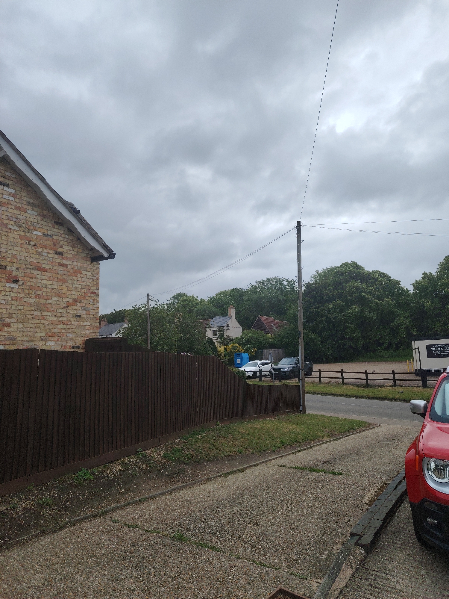 The image captures an overcast daytime scene in a village or residential area. In the foreground on the left, a dark brown wooden fence runs horizontally, partially obscuring the lower section of a light-colored brick house with a white fascia and dark roof. A concrete driveway and patch of grass are visible in front of the fence. On the far right, the front portion of a red car, featuring a distinct headlight and silver trim, is partially visible, parked on a drive.
In the midground, a tall utility pole with wires stands prominently, with wires extending across the cloudy sky. A narrow paved road runs across the frame, bordered by a low wooden post-and-rail fence on the right. Behind this fence, a parking area contains several vehicles: a white car, a dark grey SUV, and a blue portable toilet-like structure. Further right in this area, a white trailer or temporary building displays prominent text: "OLD WESTON VILLAGE HALL," with smaller text below reading "Serving the community since 1957" and "available for hire."
The background features a dense line of mature green trees, with several houses, some featuring dark or red-tiled roofs, partially visible through the foliage. The sky is uniformly covered with thick, grey clouds, suggesting an overcast day. No people are present, and no particular activity is taking place, presenting a quiet, static view.