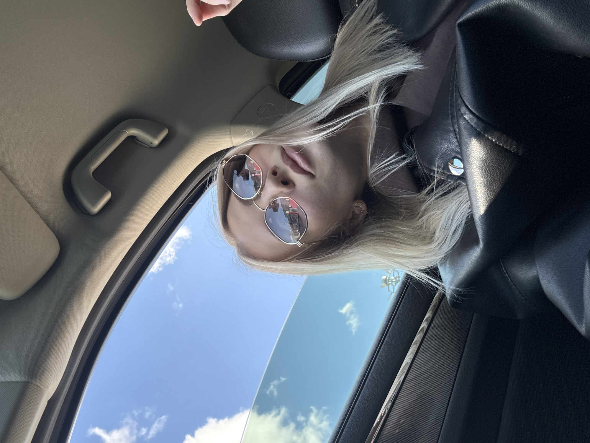 A woman with blonde hair and sunglasses is in a car, the open roof reveals a blue sky with clouds. The reflections in the sunglasses show buildings which could be the city of Izhevsk, Russia. The interior of the car shows the grab handle on the ceiling and the details of the seat and side panel. The woman is wearing a black leather jacket.