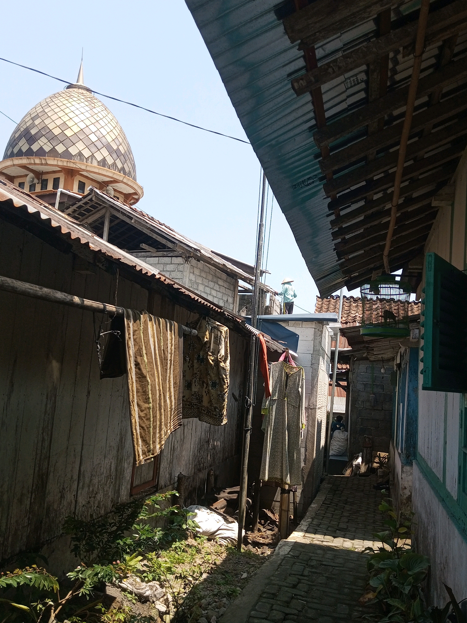 This image captures a narrow alleyway between buildings, likely in a residential or mixed-use area of Andongsili, Indonesia. The alley is paved with cobblestones and illuminated by sunlight, casting shadows. On the left, a weathered wooden wall features laundry hanging on a pole, including striped and patterned fabrics. Further back, a distinctive mosque dome with a checkered pattern rises above the rooftops, topped by a spire.
To the right, buildings with corrugated metal roofs and visible wooden beams create an overhang, partially obscuring the sky. A person wearing a conical hat stands on a rooftop, looking into the distance. Another building on the far right has a green-framed window. The scene suggests a humid, possibly tropical climate, with abundant greenery in the foreground. Clotheslines and makeshift structures indicate a lived-in environment. The overall impression is one of a dense, possibly older urban or semi-urban settlement.