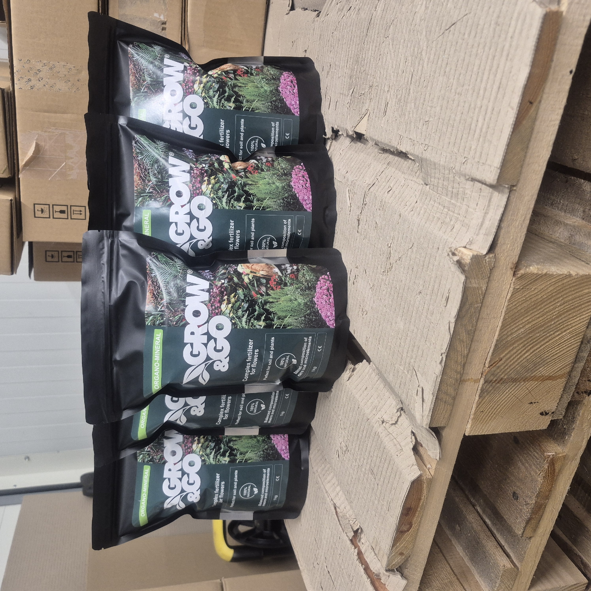 The image shows a stack of black pouches of fertilizer, labeled "GROW & GO ORGANO-MINERAL". The pouches are positioned vertically, with the text and imagery oriented sideways. Each pouch features a colorful illustration of flowers and plants, and text indicating that it is a "Complex fertilizer for flowers", "Perfect for soil and plants", "100% NATURAL", and contains a "Balanced composition of macro and microelements" in a "1kg" package. The CE mark is also visible on the pouches. The fertilizer bags are resting on a wooden pallet, which appears weathered and rough. In the background, cardboard boxes are stacked, suggesting a storage or warehouse setting. The lighting suggests an indoor environment, possibly a warehouse or distribution center in Nowa Wieś Książęca, Poland. No people are visible in the image.