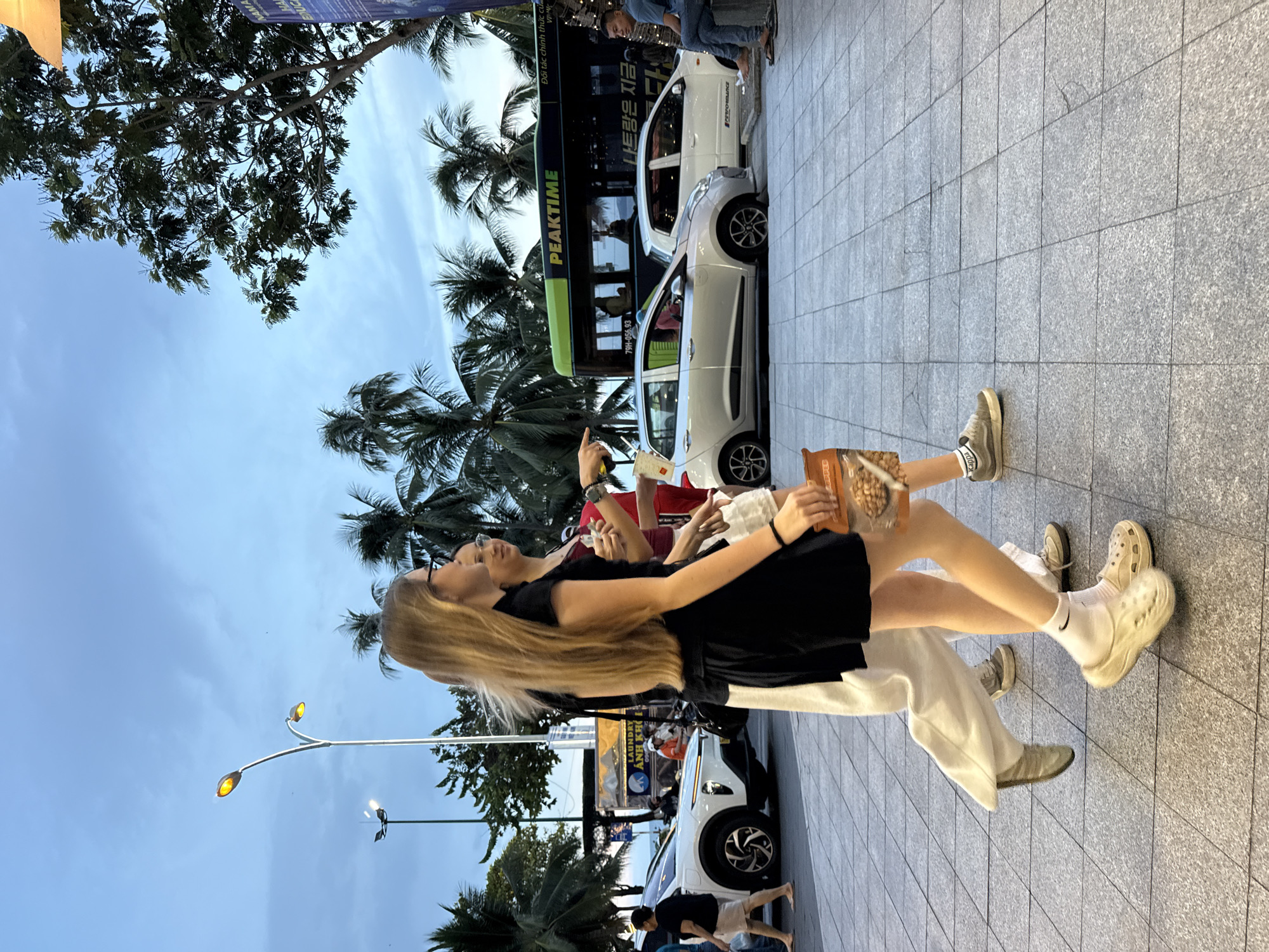 The image depicts a street scene in Nha Trang Ward, Vietnam, during twilight. Two young women are walking on a tiled sidewalk. The woman in the foreground is wearing a black short-sleeved dress, white socks, and beige clogs. She has long blonde hair and is holding a bag of snacks. The woman behind her is wearing a red top and a white skirt, and she appears to be gesturing with her hand. Behind them, several cars are parked, including a silver hatchback and a white sedan. A green and black bus with "PEAKTIME" written on its side is also visible. Palm trees line the street, and streetlights illuminate the scene. In the background, a man is sitting on a ledge. The sky is overcast with hues of blue and grey.