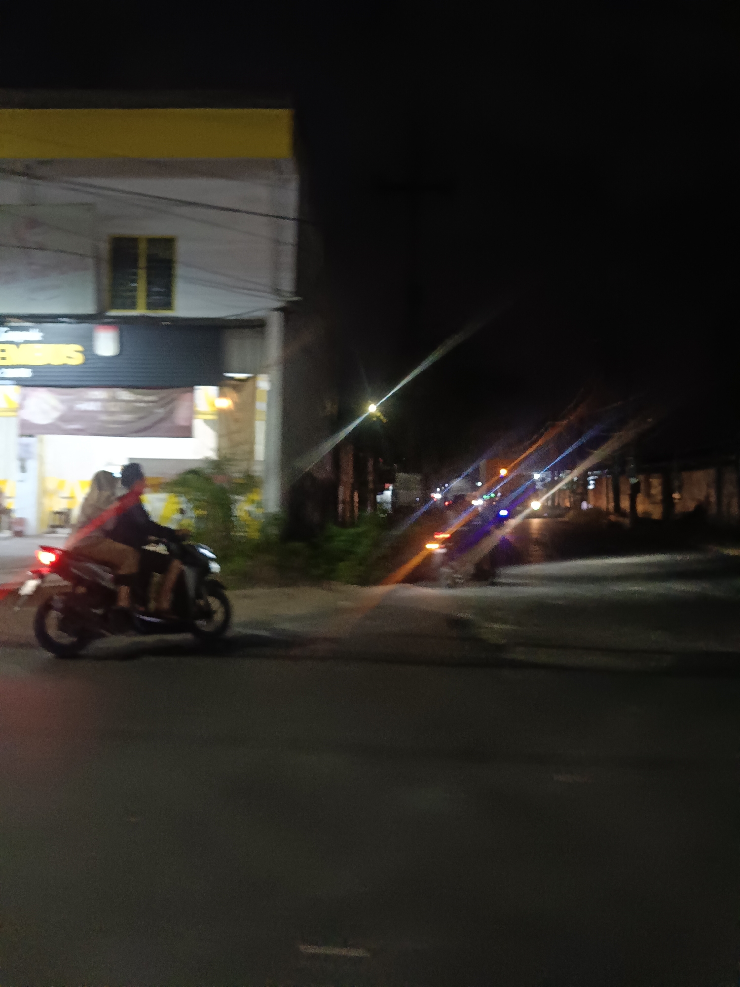 The image depicts a nighttime scene on a street in Tropodo, Indonesia. In the foreground, two individuals are riding a scooter. The person in front is wearing a dark blue long-sleeved shirt and a dark hat, while the person behind them is wearing a light-colored headscarf and a light-colored jacket. The scooter has a red taillight illuminated. To the left of the scooter, a building with a yellow awning and a sign that reads "PEMBUS" is visible. The building has a white facade and a window with yellow panes. The background is dark, with streaks of light suggesting other vehicles or streetlights. The street is paved, and there is vegetation on the right side of the image.